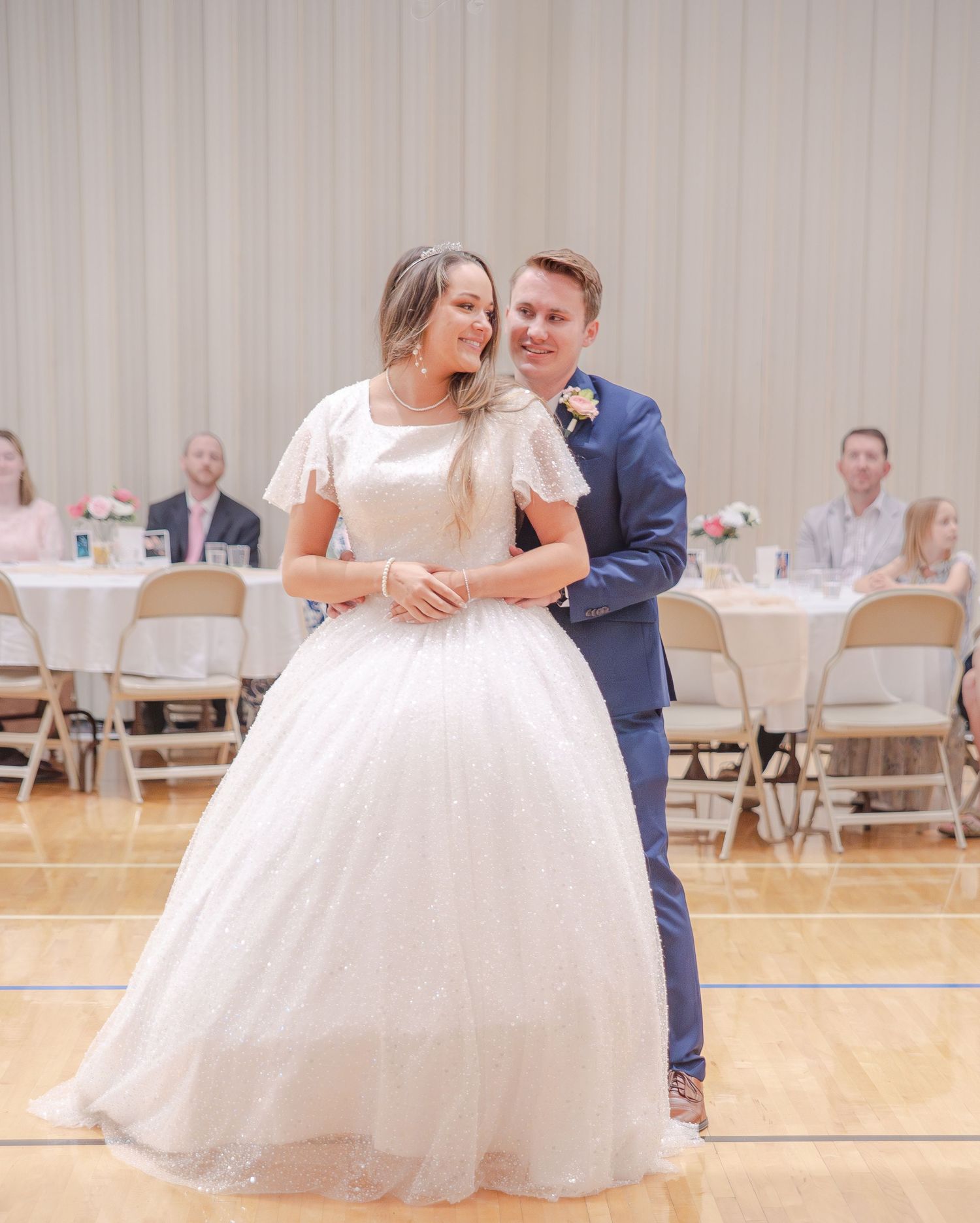 A couple shares their first dance at their wedding reception in a gymnasium with white chairs in the background.