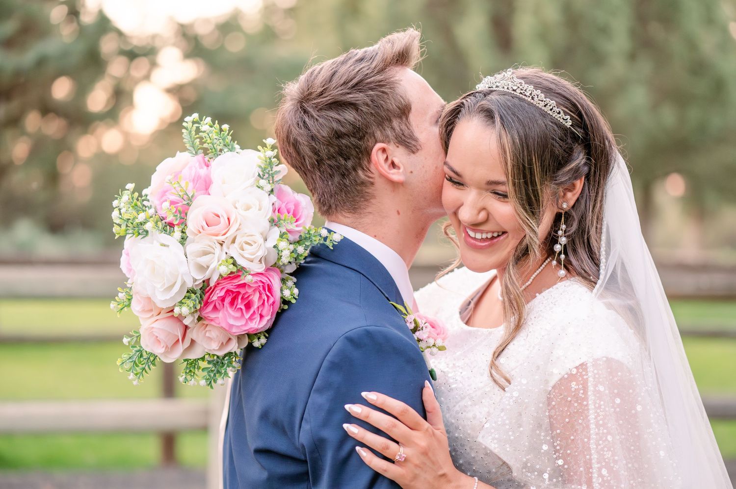 Romantic outdoor wedding couple embraces in garden setting with pink roses and white lace dress details.