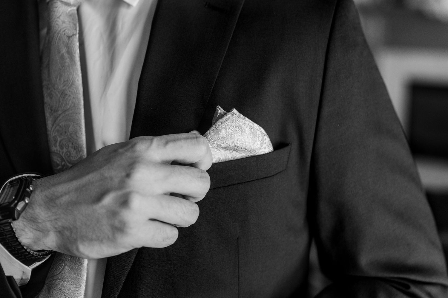 Black and white photo of a groom in a dark suit placing a white pocket square into his breast pocket.