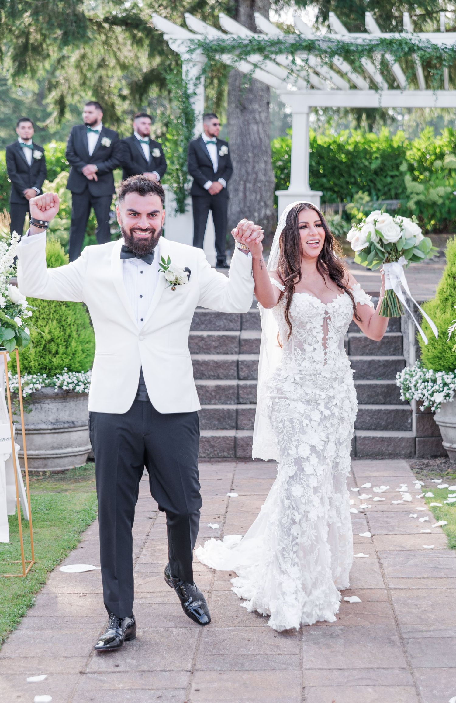 Joyful newlyweds celebrate after outdoor wedding ceremony, bride in lace gown holding bouquet, groom in white tuxedo jacket.