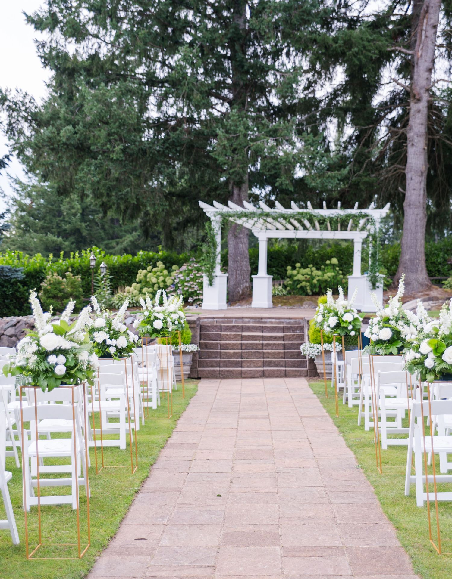Elegant outdoor wedding ceremony aisle with white chairs, floral arrangements, and a white pergola altar surrounded by tall trees.