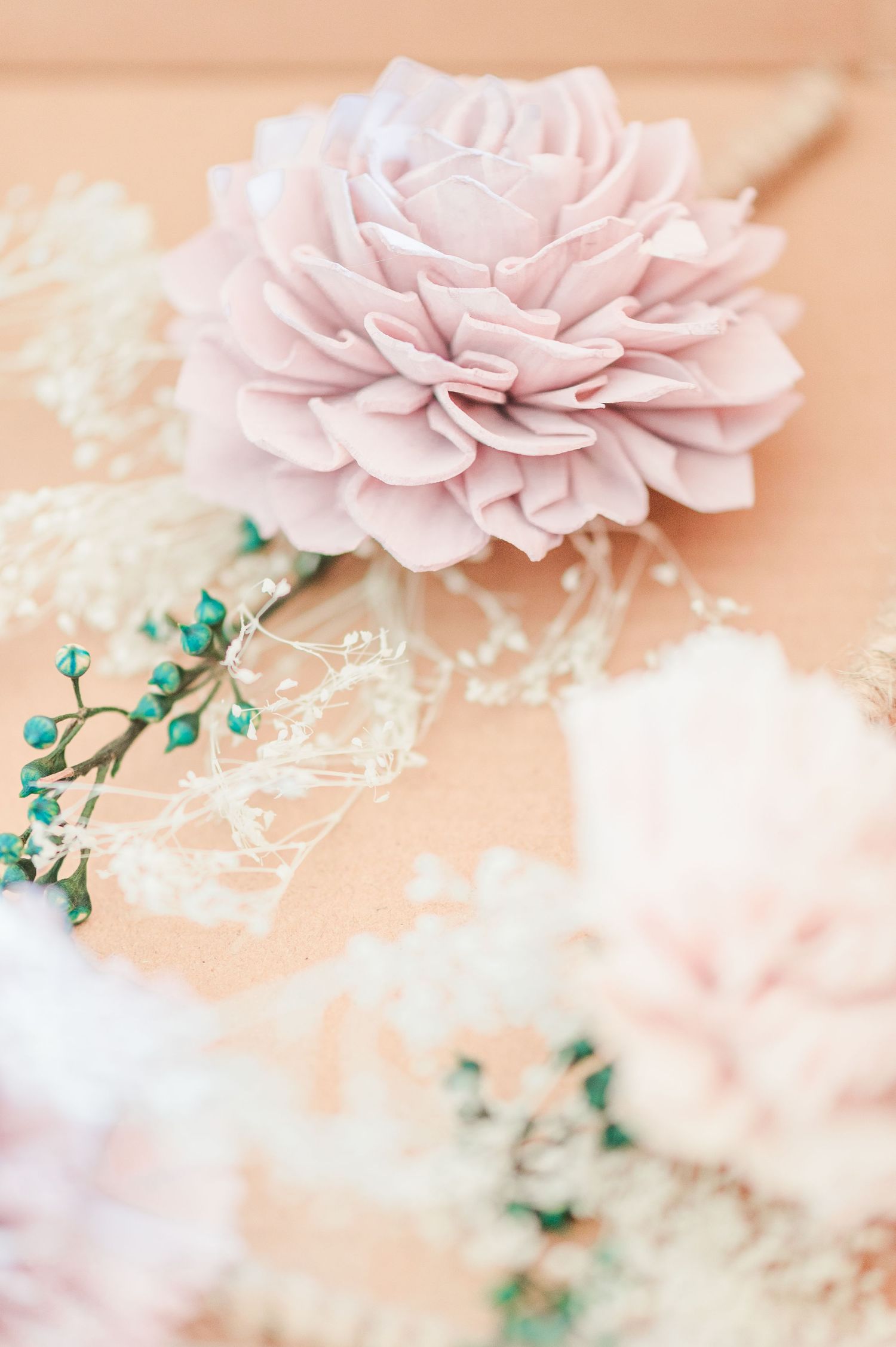 Soft pink fabric flower with delicate white baby's breath and teal berries on a kraft paper background.