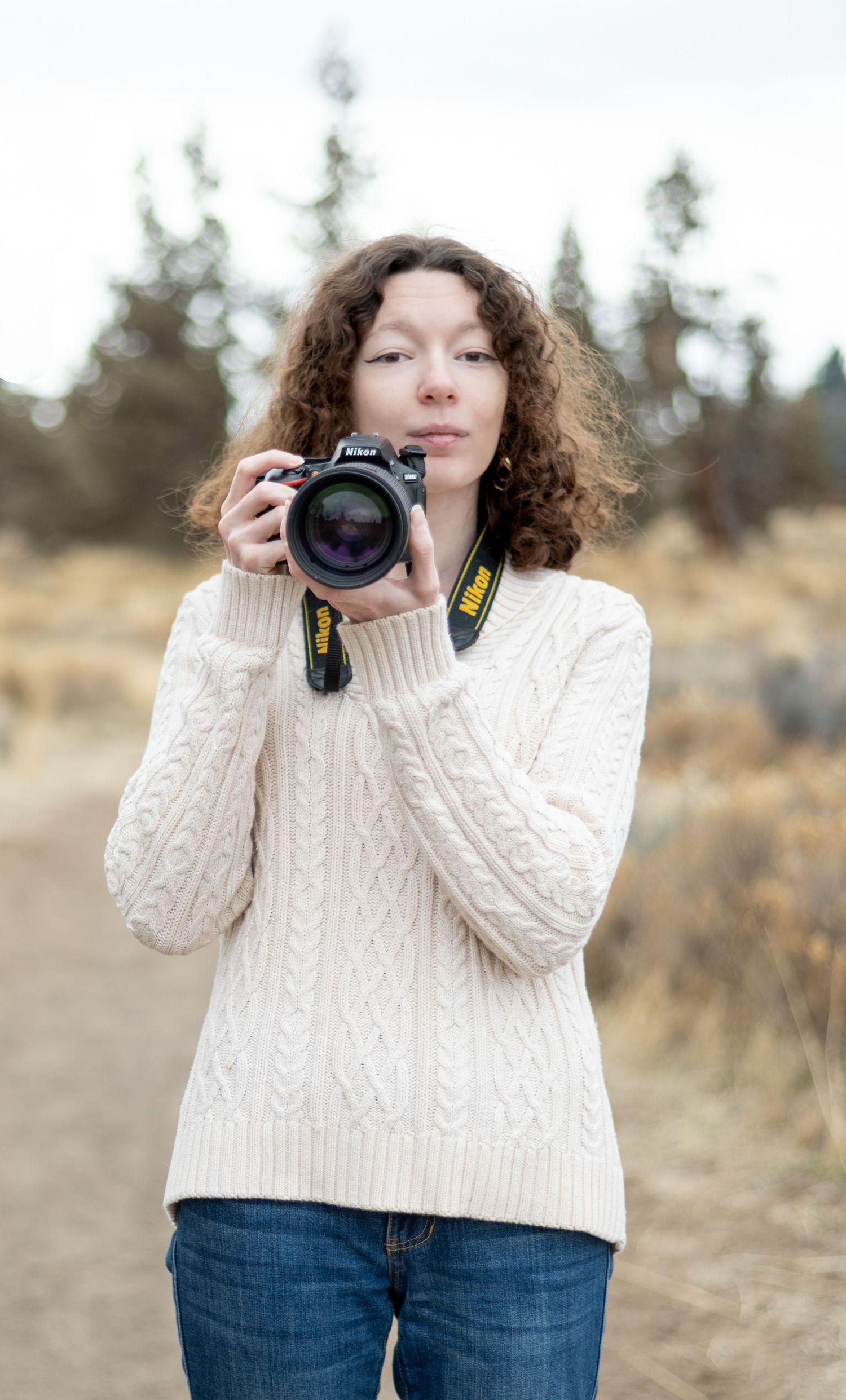Photographer in white cable knit sweater demonstrating camera poses in outdoor setting.