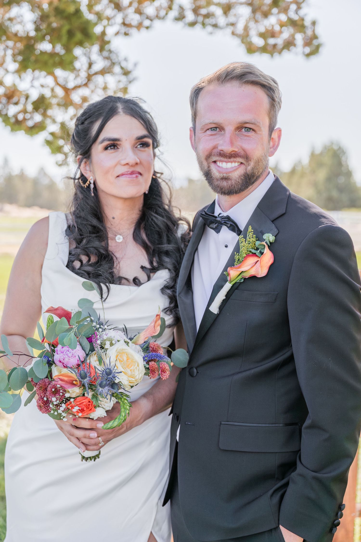A smiling bride holding a colorful bouquet stands beside a groom in a dark suit with boutonniere at an outdoor wedding.