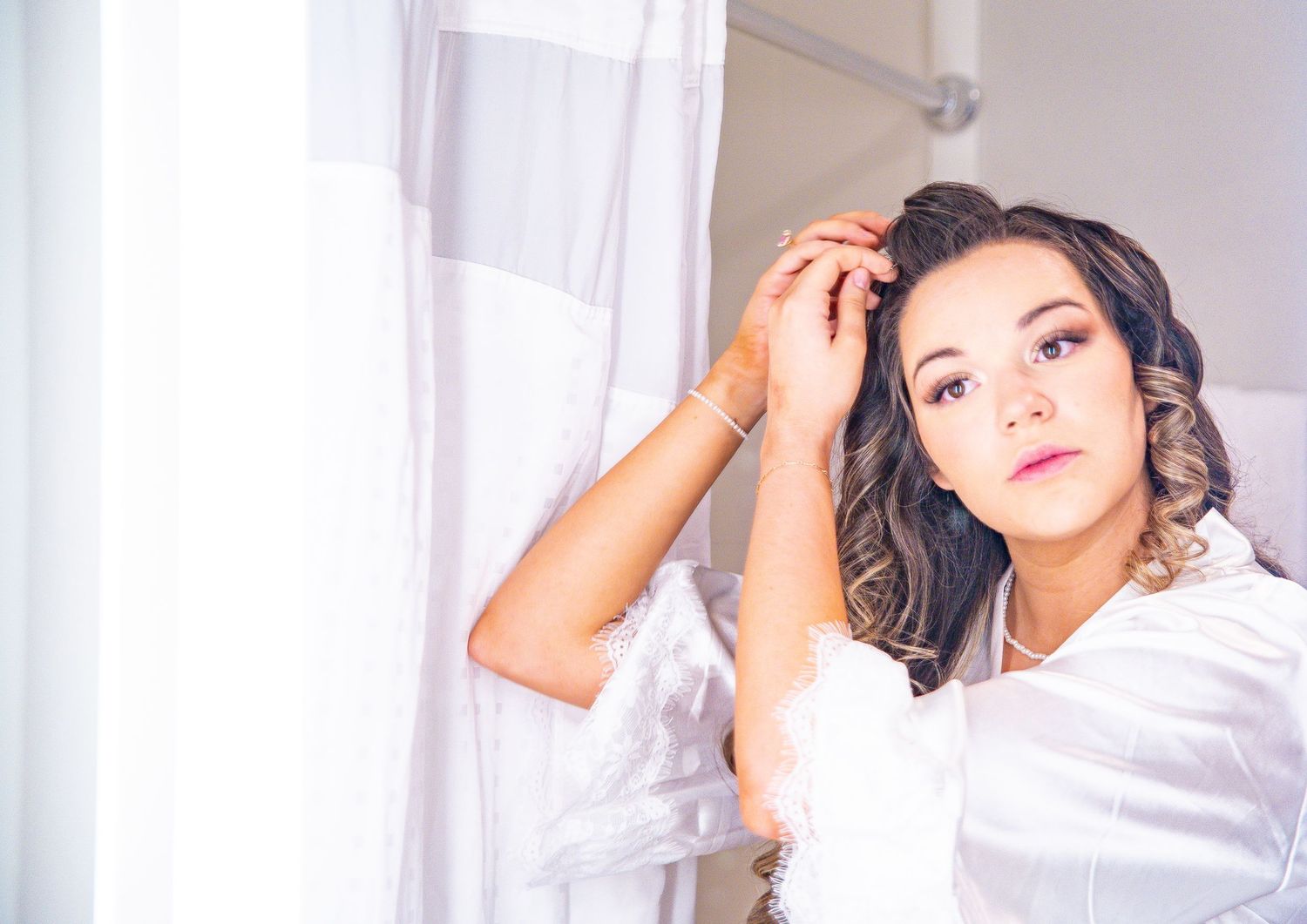 Bride in white robe styles her hair during wedding day preparations.