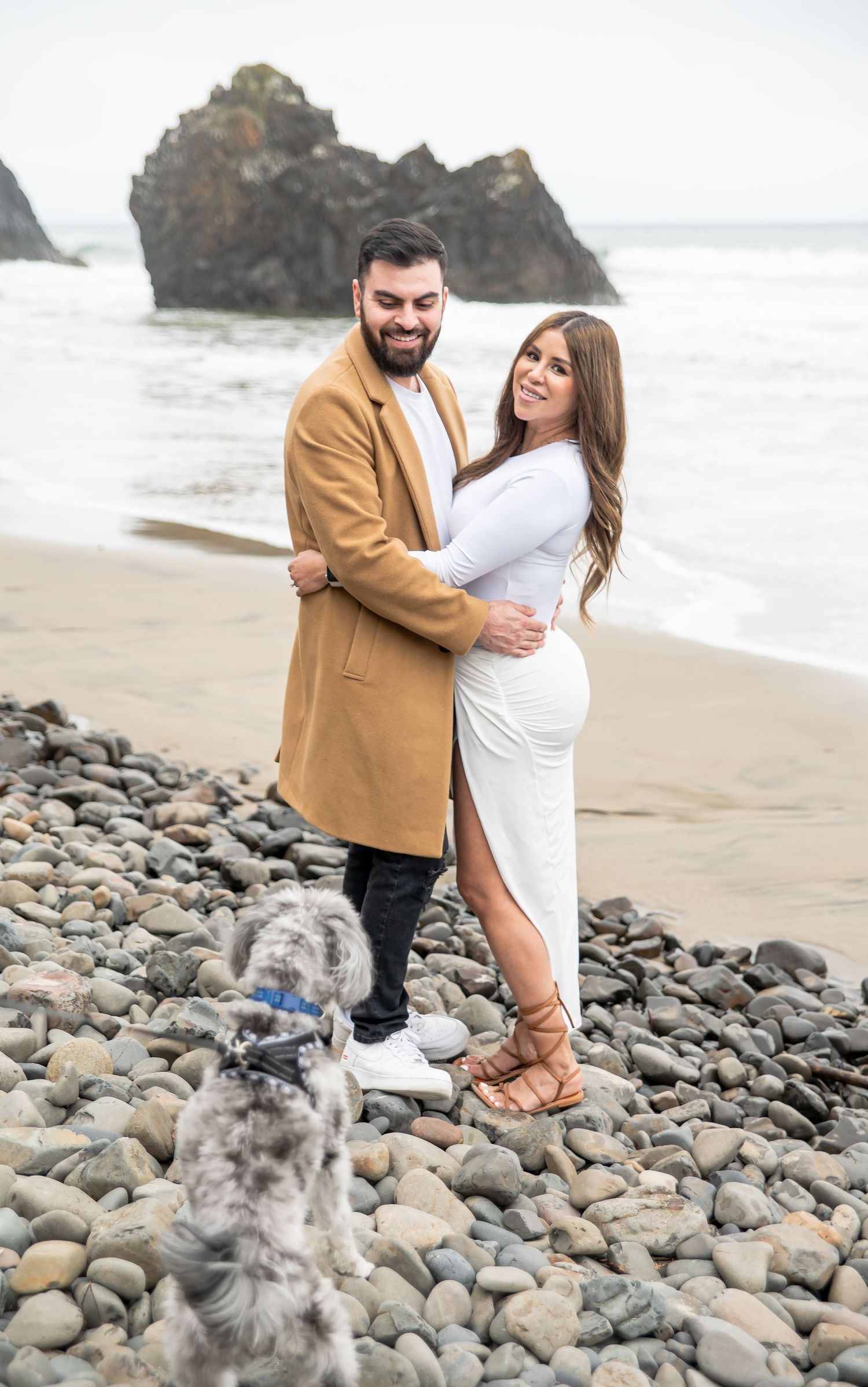 Couple embracing on rocky beach with dog, woman in white dress, man in tan coat, ocean waves and rock formation behind them.