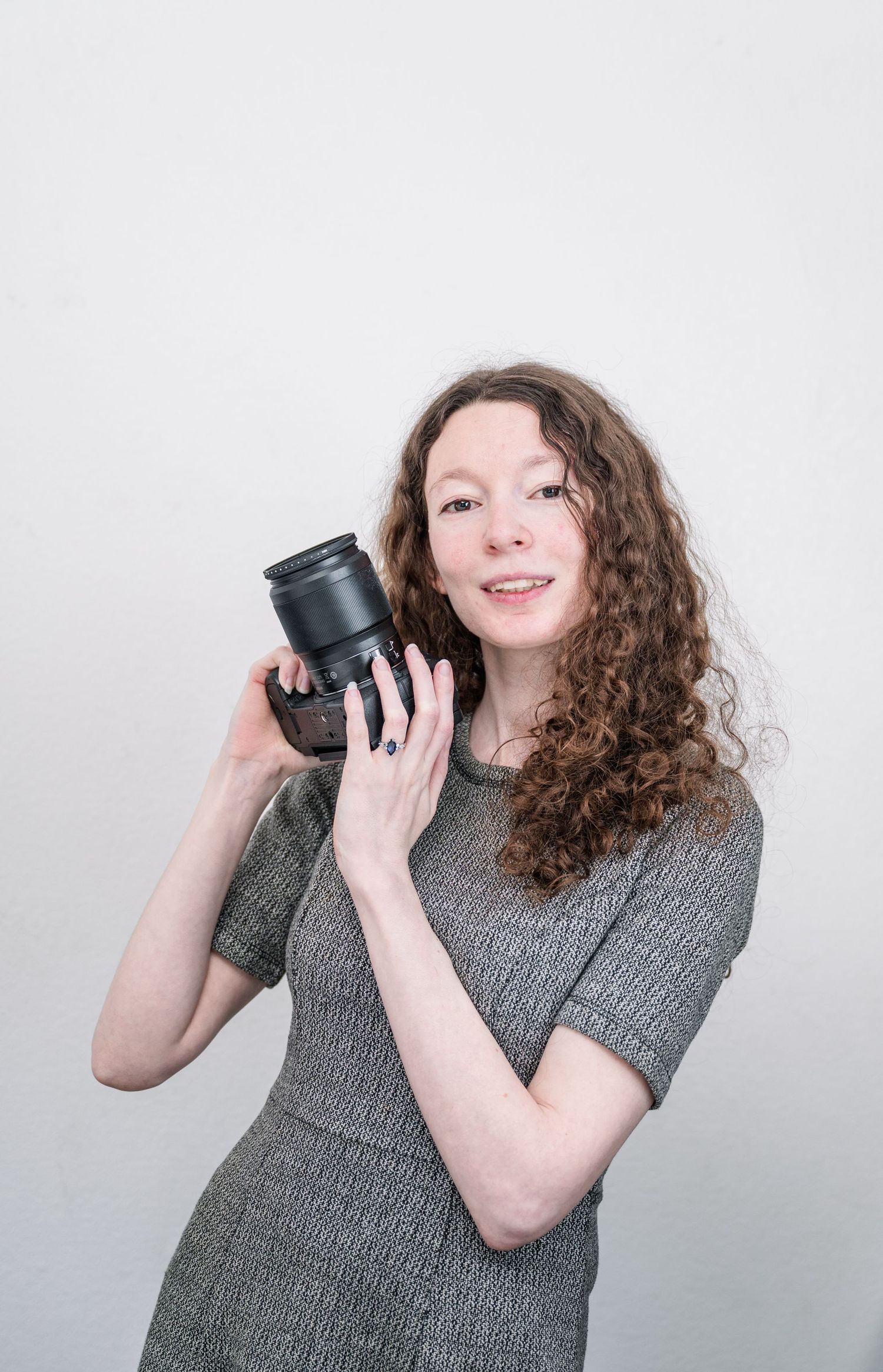 Young woman with curly hair holding a DSLR camera, wearing a gray dress, posing against a white background.