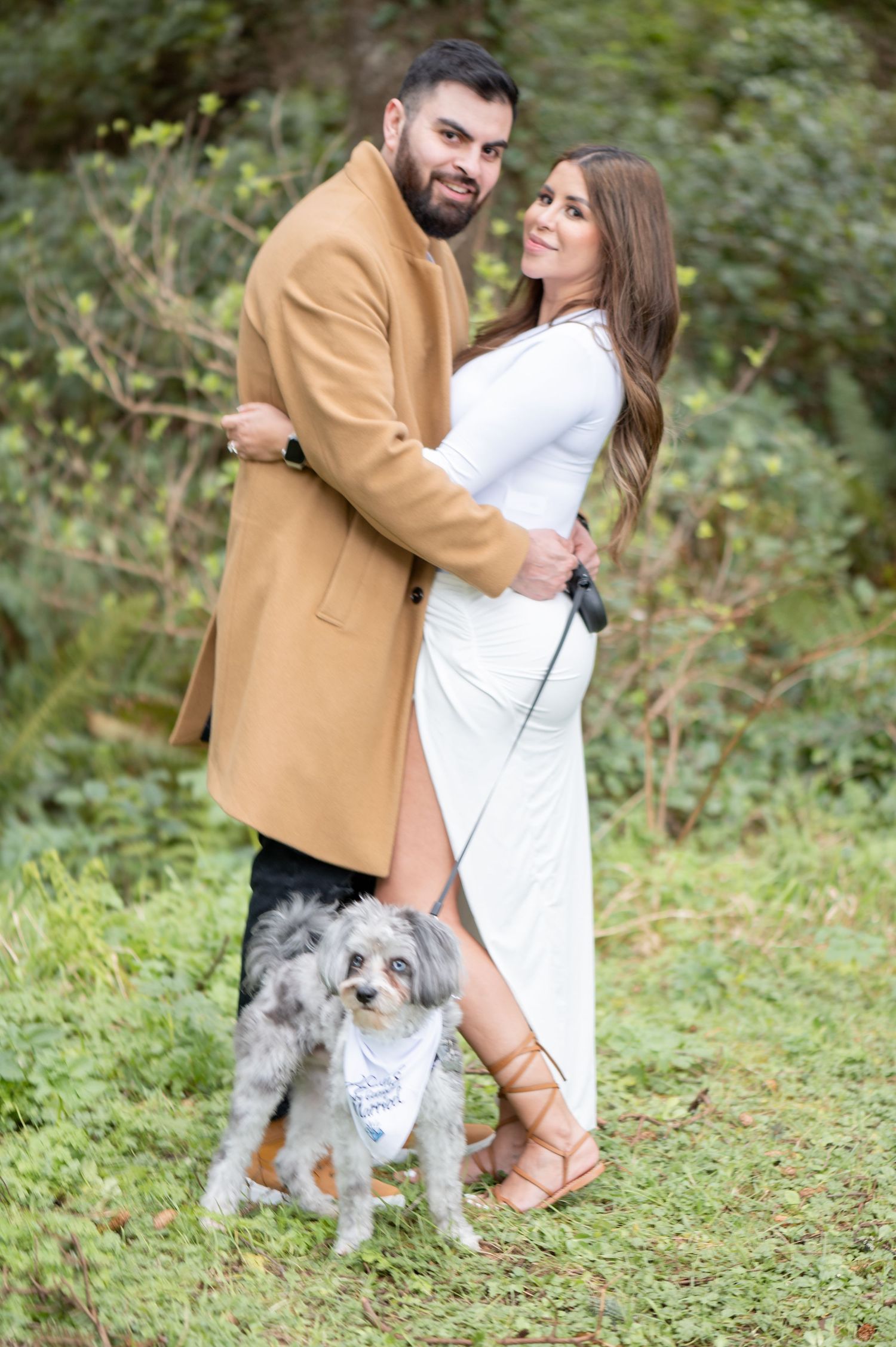 Couple in white dress and tan coat embracing outdoors with small fluffy dog on leash in green nature setting.