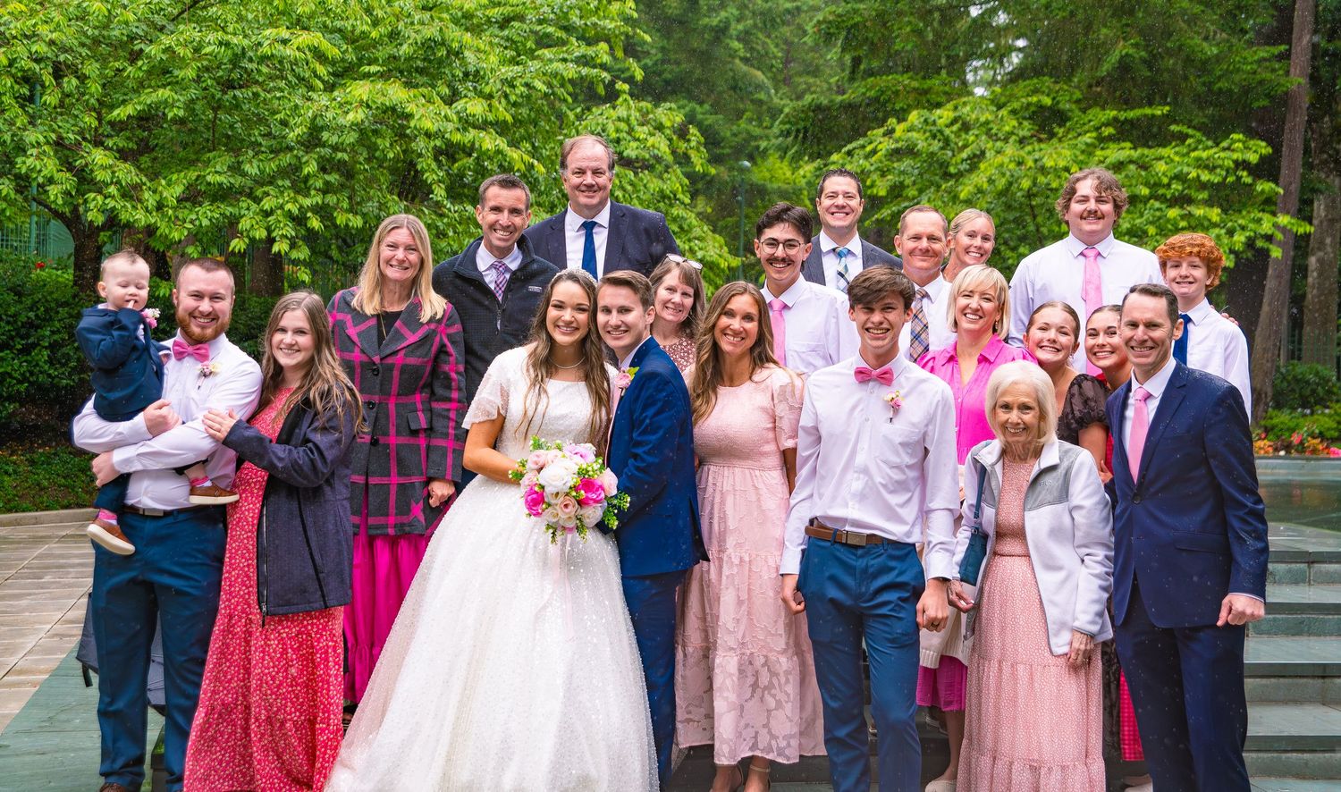 Wedding party group photo with bride in white dress and guests in pink and red dresses against green garden backdrop.