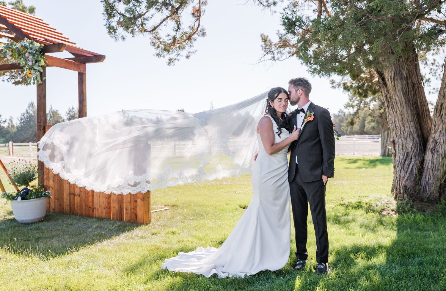Bride and groom share a romantic moment outdoors, her veil flowing in the wind, with a wooden pergola and green trees behind.