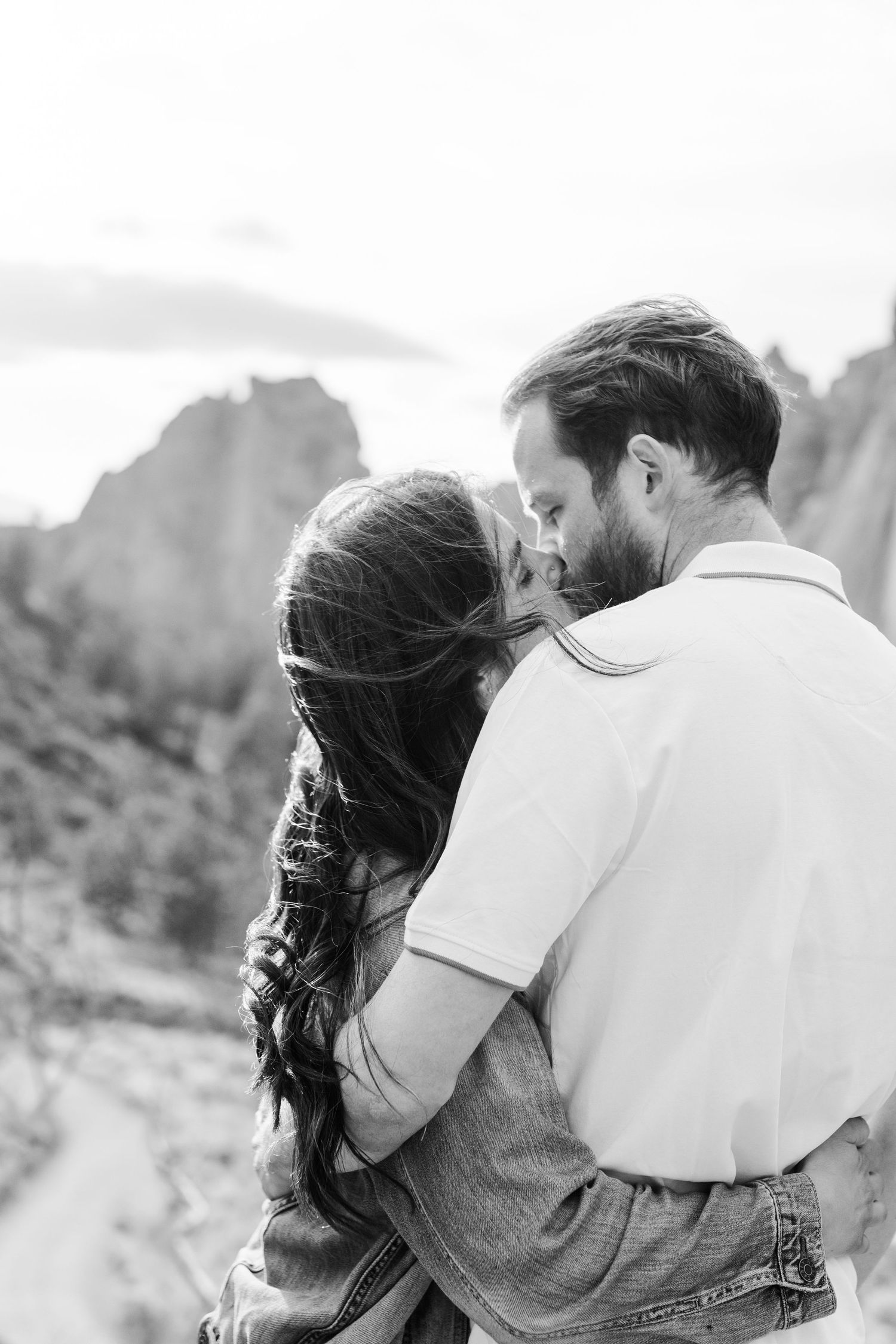 Black and white photo of a couple embracing and almost kissing outdoors with rocky cliffs in the background.