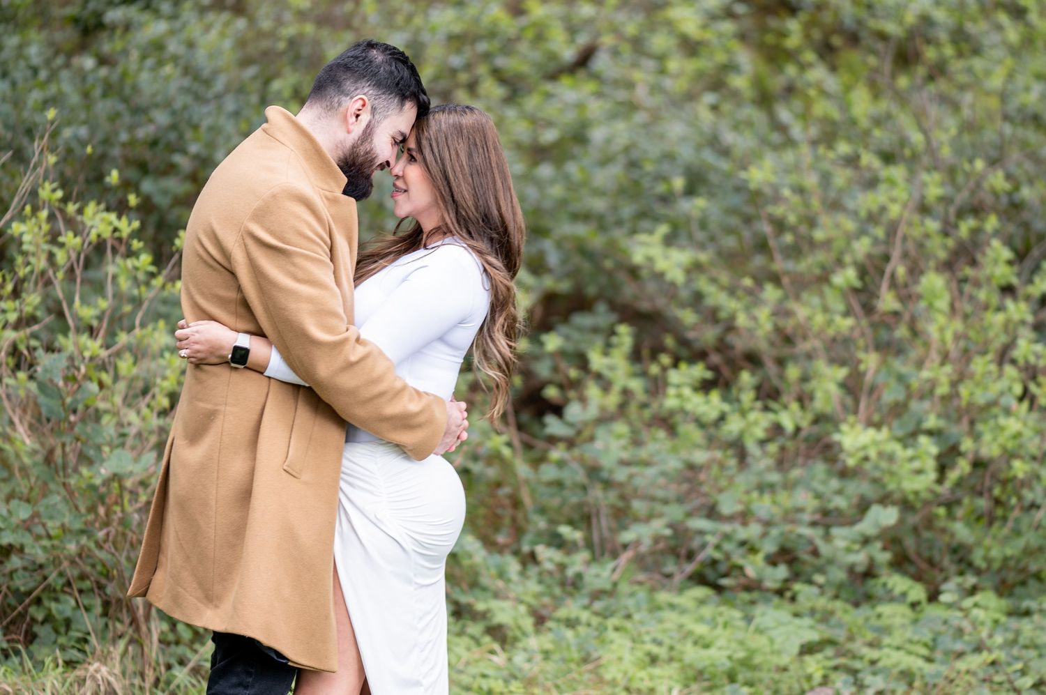Couple sharing a tender moment outdoors, pregnant woman in white dress embraced by man in tan blazer amid lush greenery.