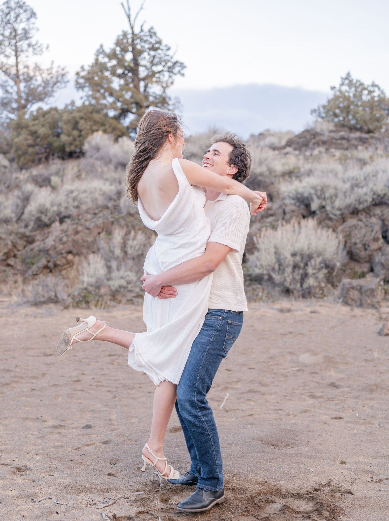 A joyful couple embraces outdoors, man lifting woman in white dress, with desert shrubs and mountains in the background.