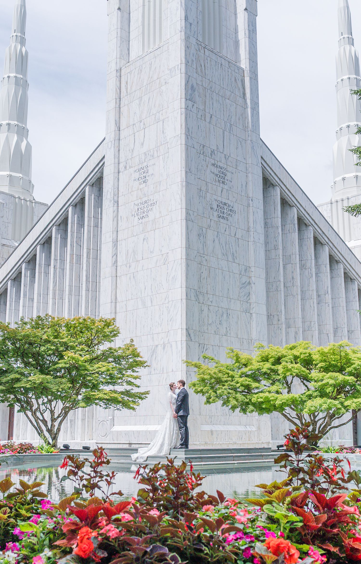 Bride and groom embrace at the base of a grand white marble temple surrounded by colorful flowers and lush green trees.