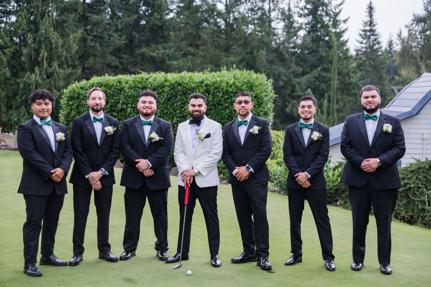 Groom in white tuxedo stands with six groomsmen in black suits and bow ties on a golf course lawn.