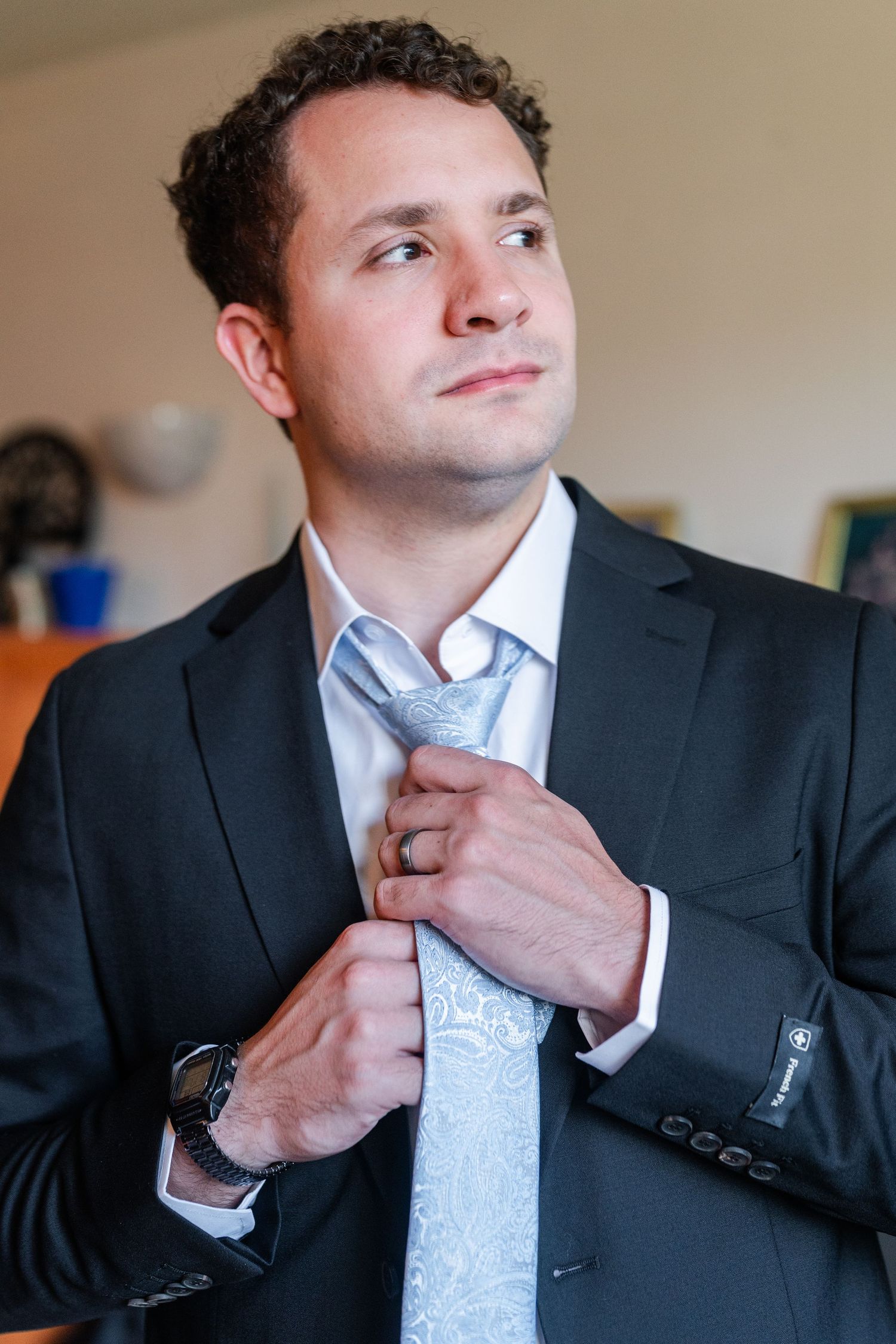 A groom in a dark suit adjusts his light blue paisley tie while looking away, wearing a watch on his wrist.
