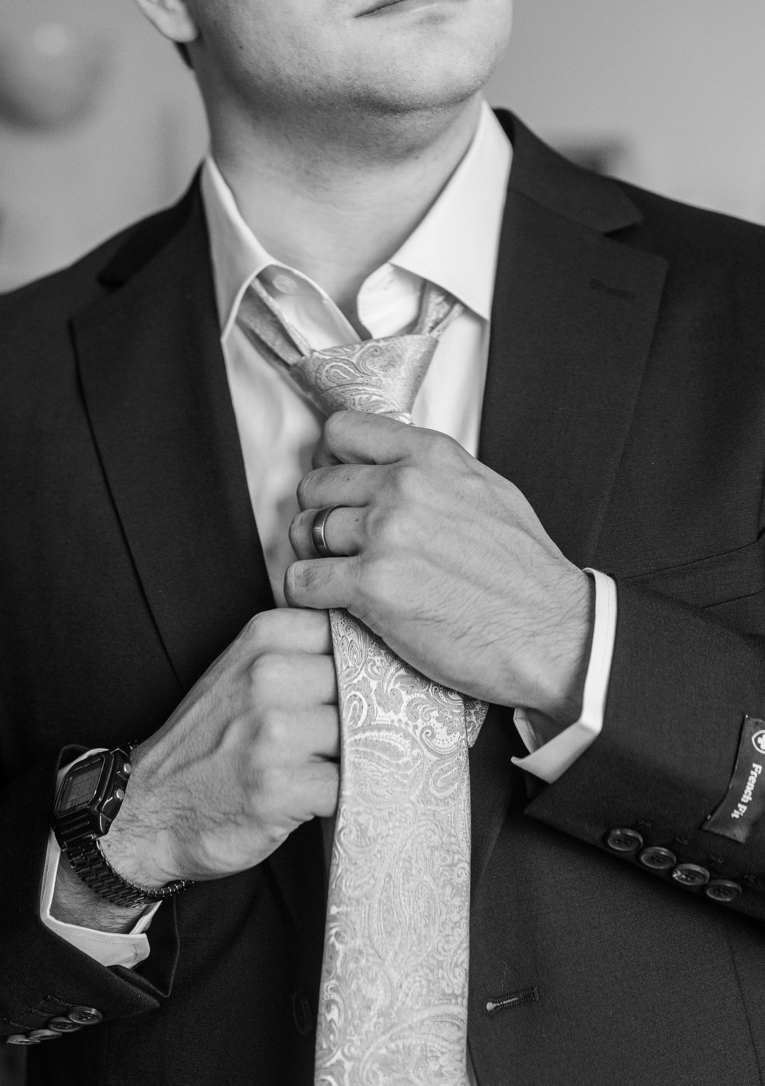 Black and white photo of a man in a dark suit adjusting his paisley tie, wearing a watch.