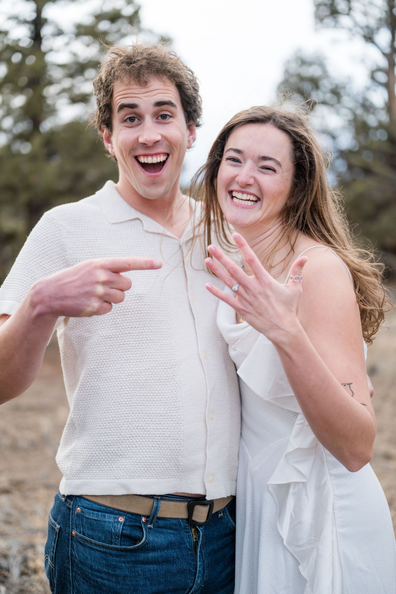 Excited couple showing off engagement ring outdoors, man pointing at ring while woman displays it, both laughing joyfully.