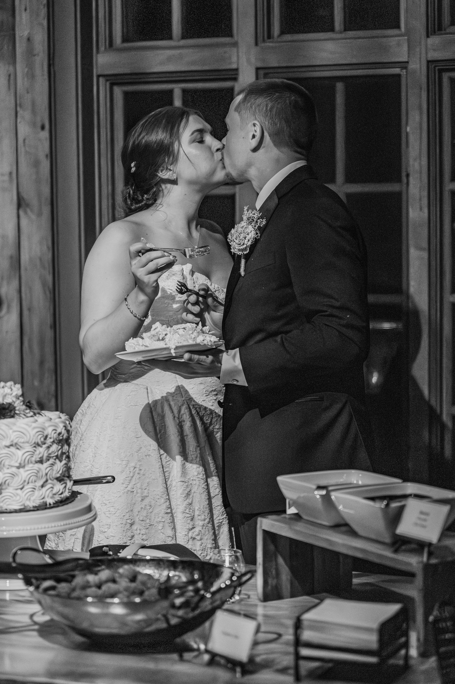 Black and white photo of a bride and groom sharing a kiss while cutting their wedding cake at their reception.
