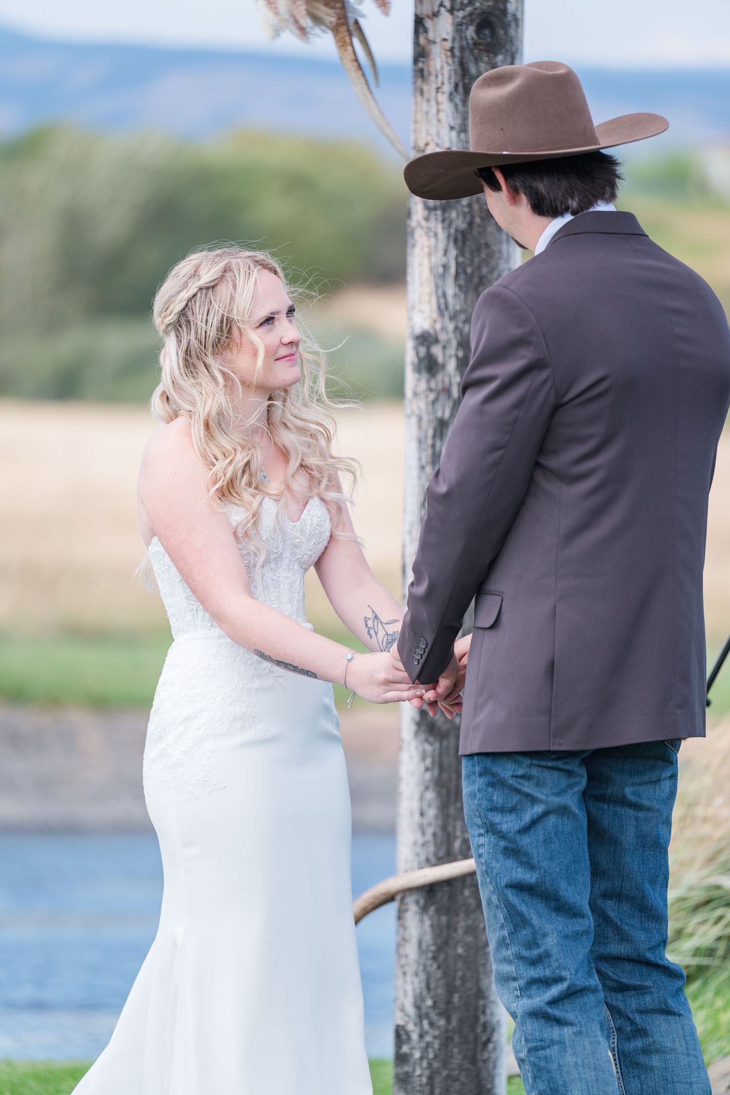 Bride in white lace gown holds hands with groom in brown blazer and cowboy hat during outdoor wedding ceremony.