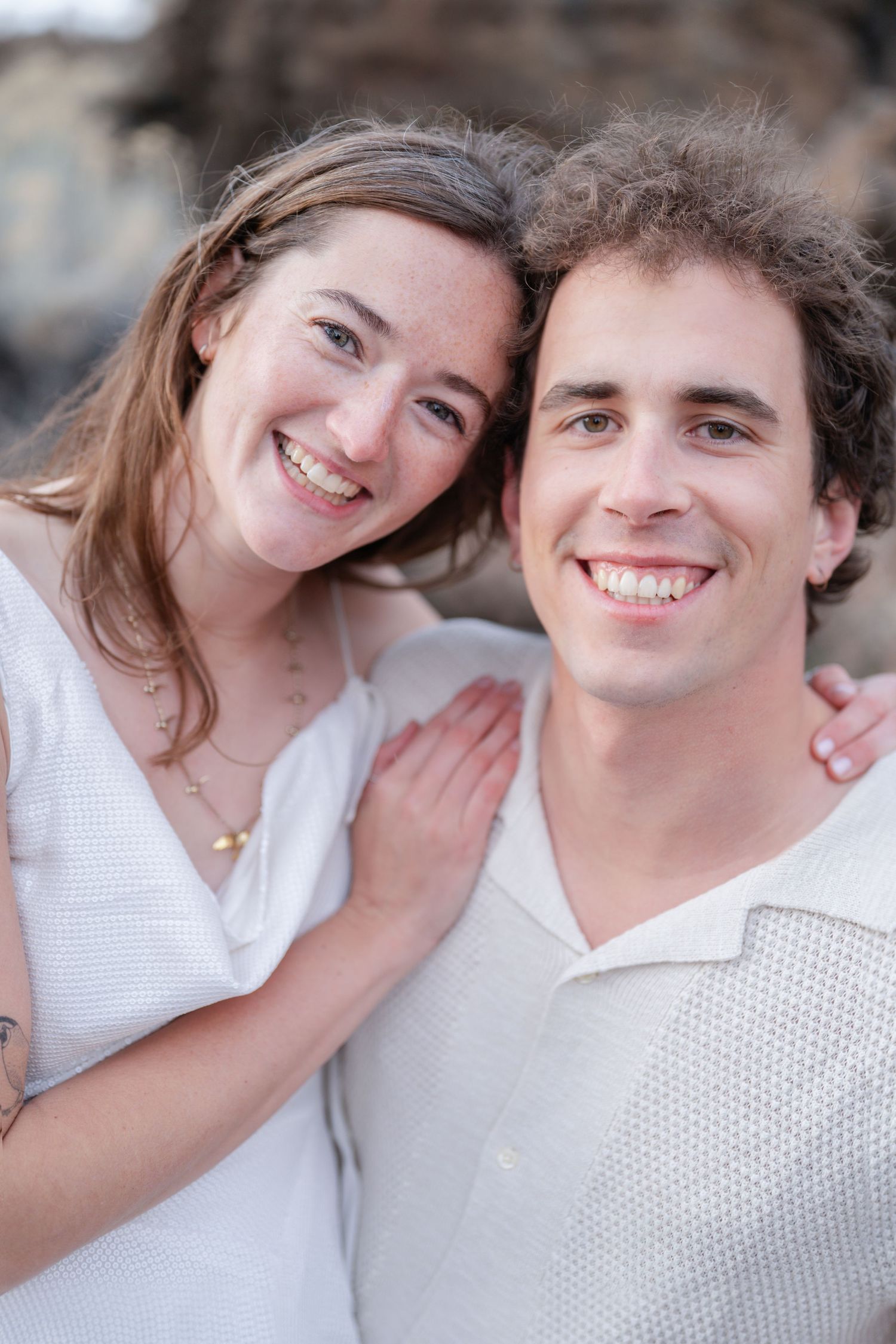A smiling young couple in white outfits posing closely together outdoors, sharing a happy moment.