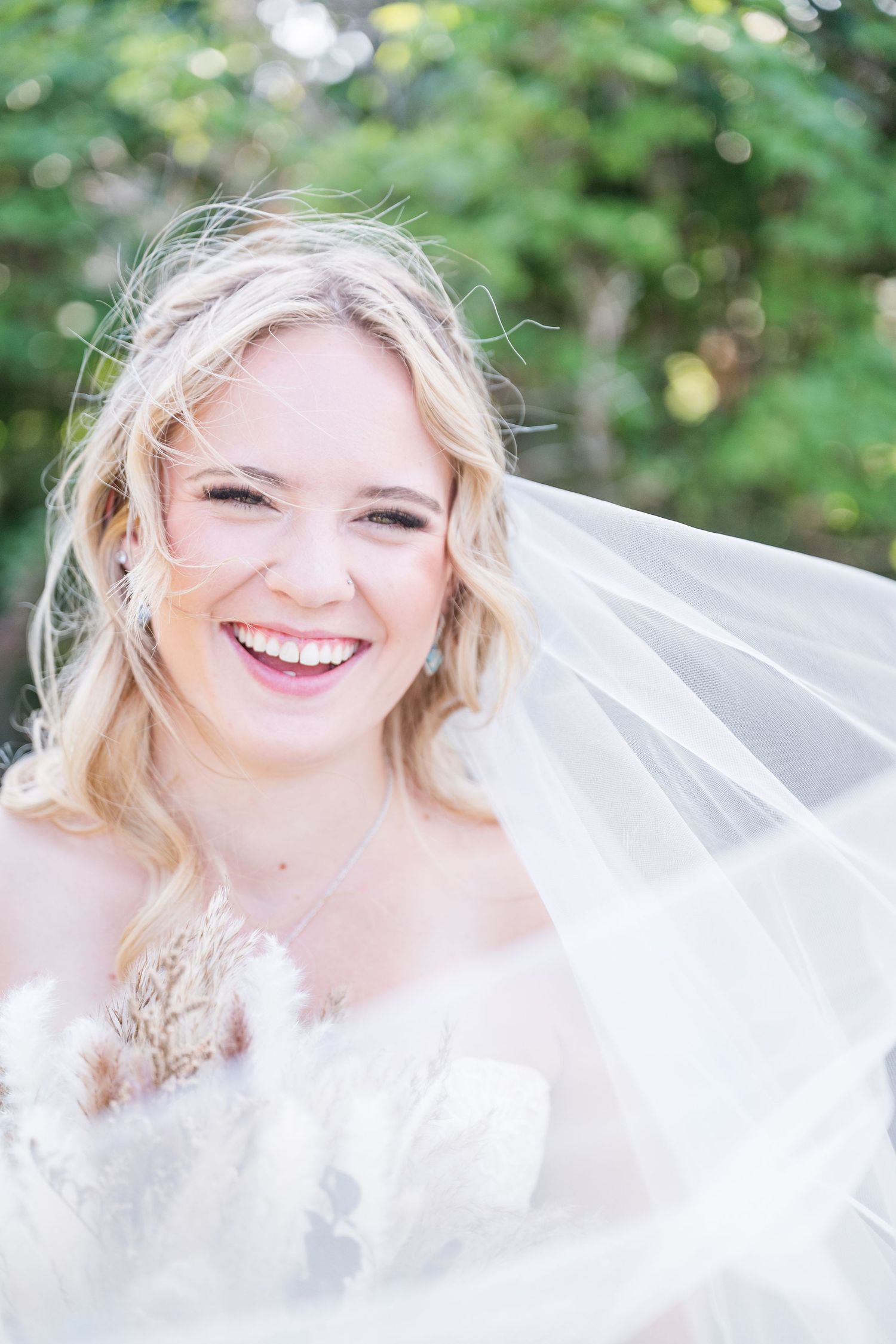 Smiling blonde bride wearing a white veil and floral gown, laughing outdoors with green trees in the background.