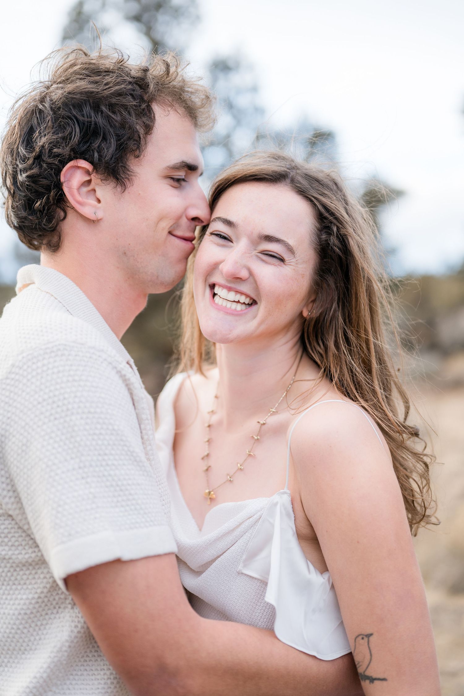 A joyful couple embraces outdoors, the man kissing the woman's cheek as she laughs, both wearing white in a natural setting.