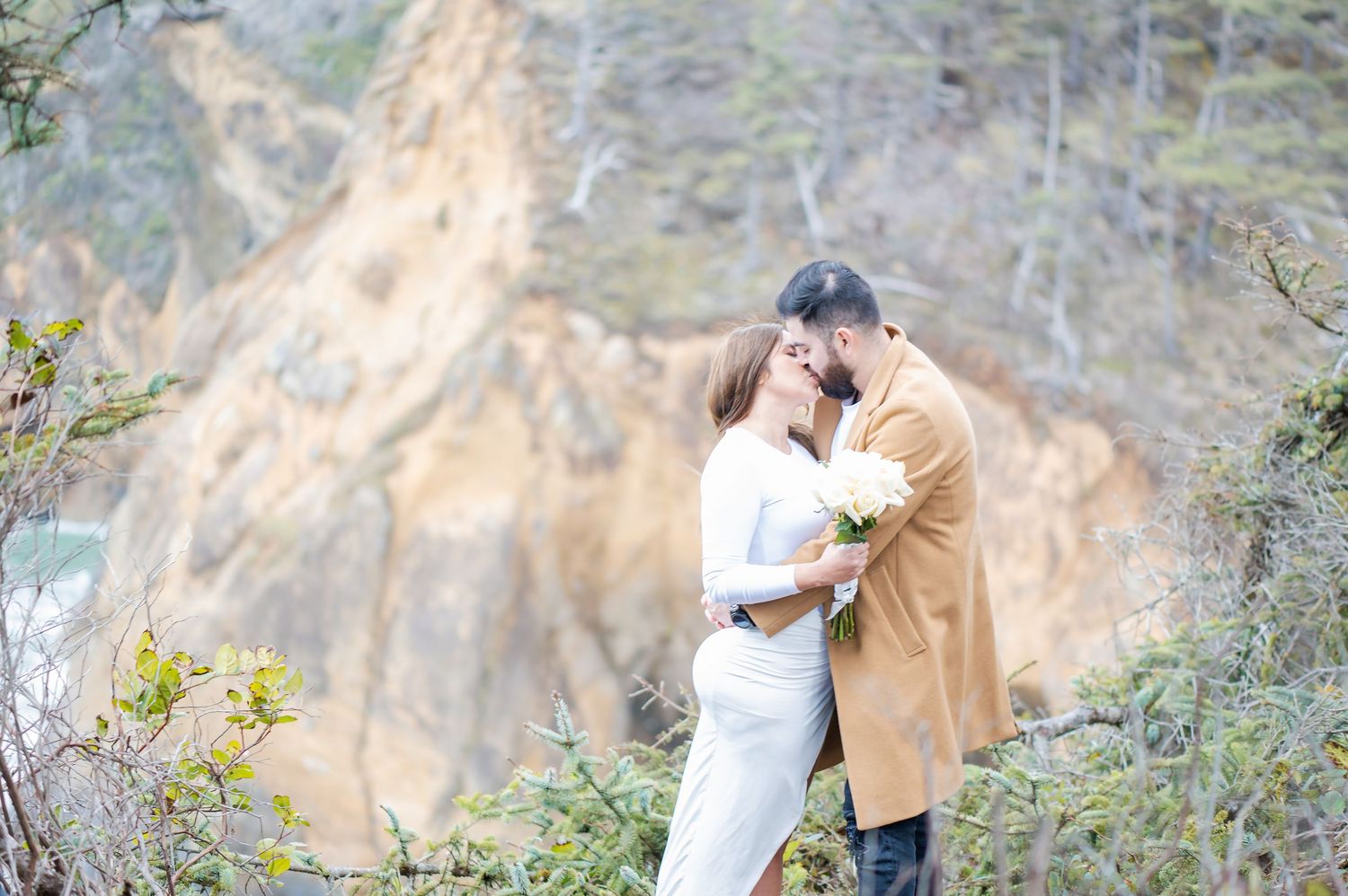 Couple sharing a romantic kiss outdoors with scenic rocky cliffs and lush greenery in the background.
