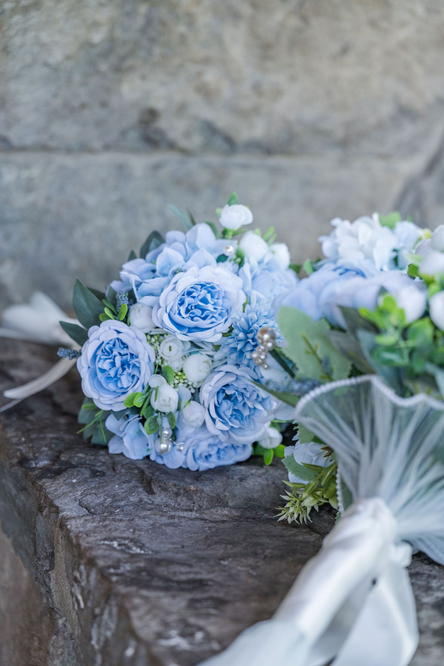 Two elegant blue and white wedding bouquets with roses and pearls resting on rustic stone steps.