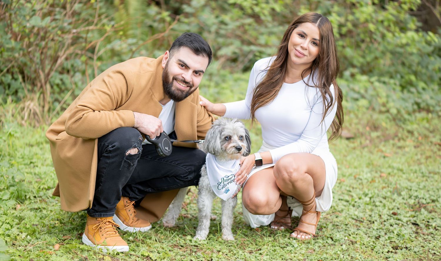 Couple crouching with small gray dog on green grass surrounded by lush trees in an outdoor photo session.