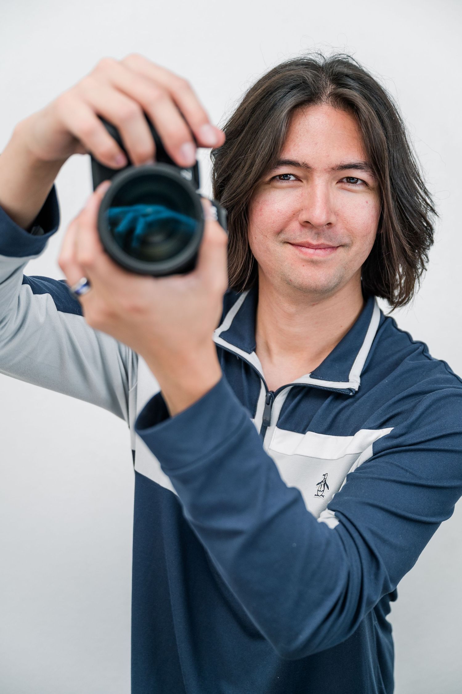 A young man with dark hair smiles while holding a DSLR camera up to shoot, wearing a blue polo shirt.