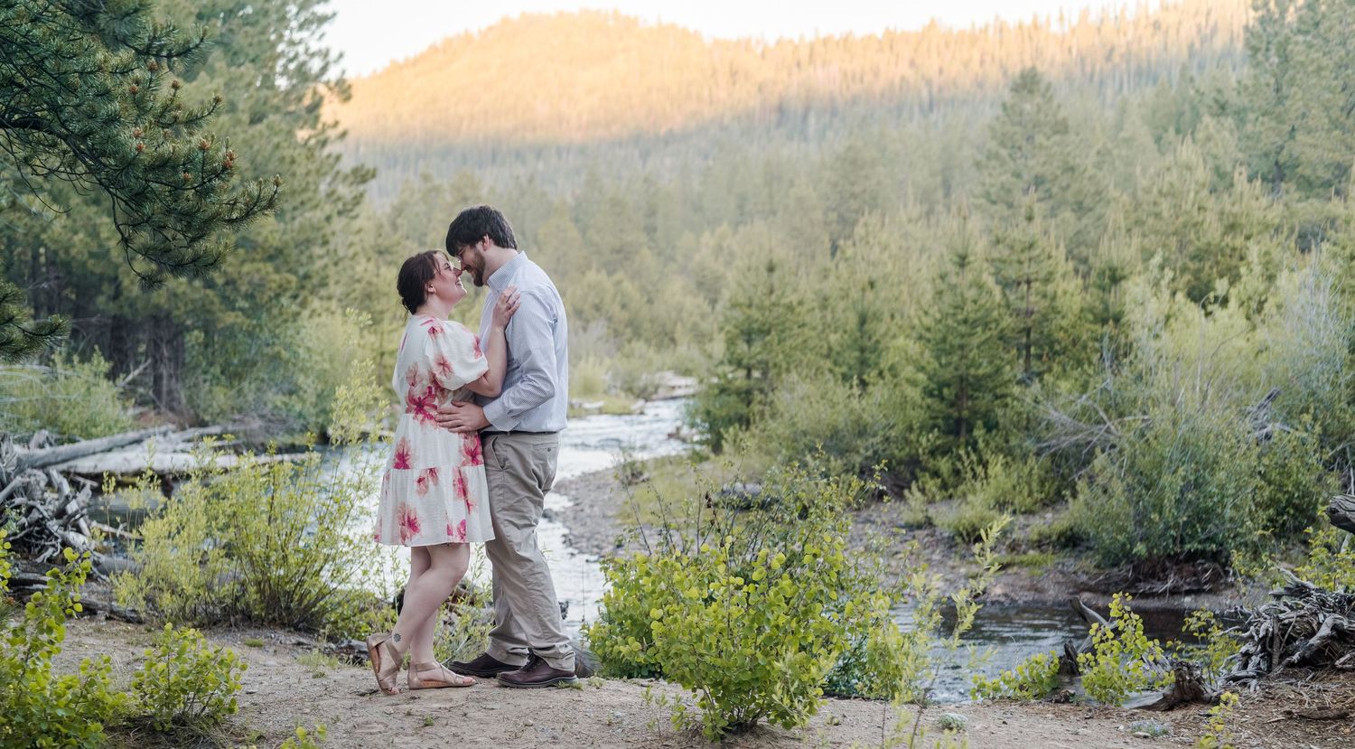 Couple sharing a romantic kiss outdoors near a mountain stream surrounded by lush green trees at sunset.