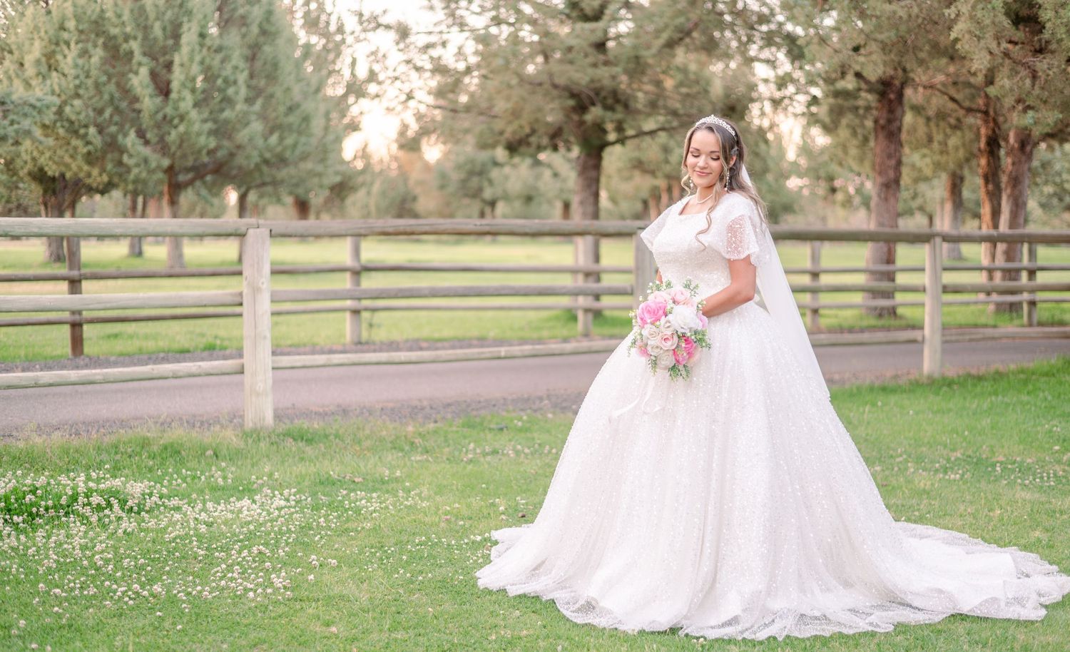 Bride in flowing white gown sits gracefully on grass near wooden fence during outdoor wedding.
