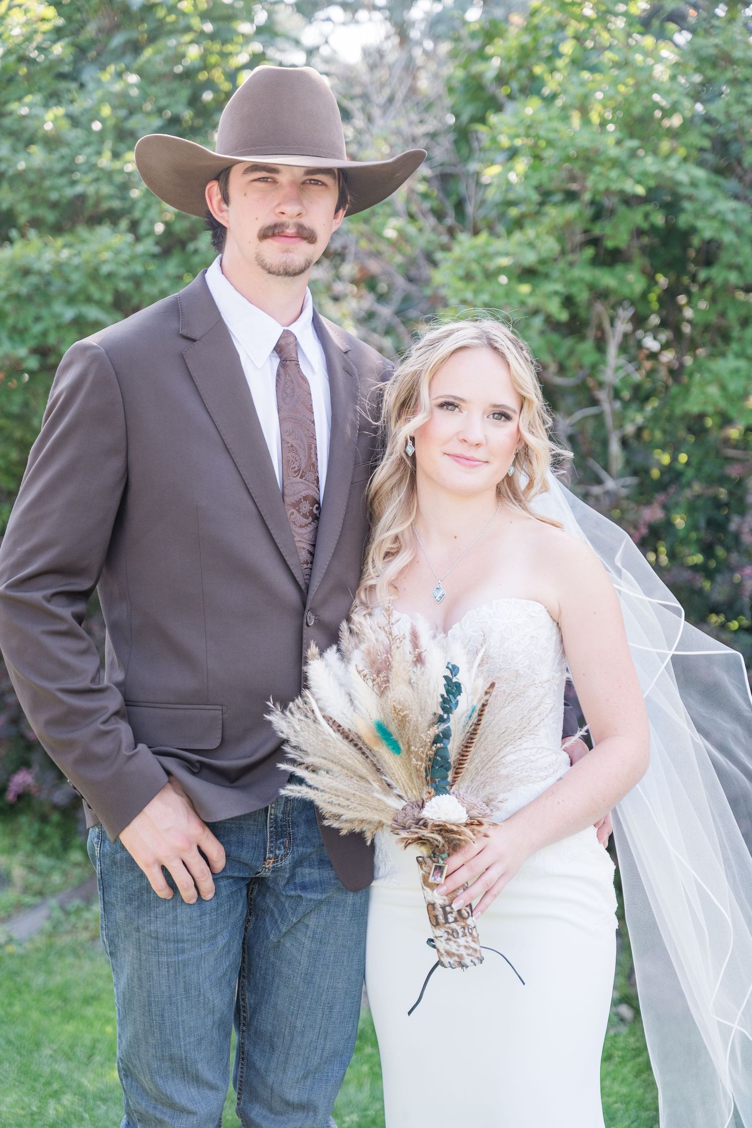 Bride and groom posing outdoors, groom in cowboy hat and brown blazer, bride in strapless gown holding boho bouquet.