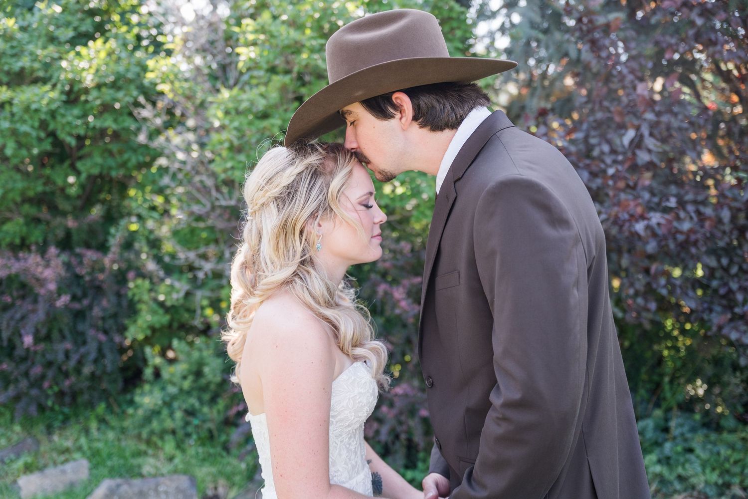 A man in a brown hat and suit gently kisses the forehead of a blonde woman in a white dress, surrounded by lush green foliage.