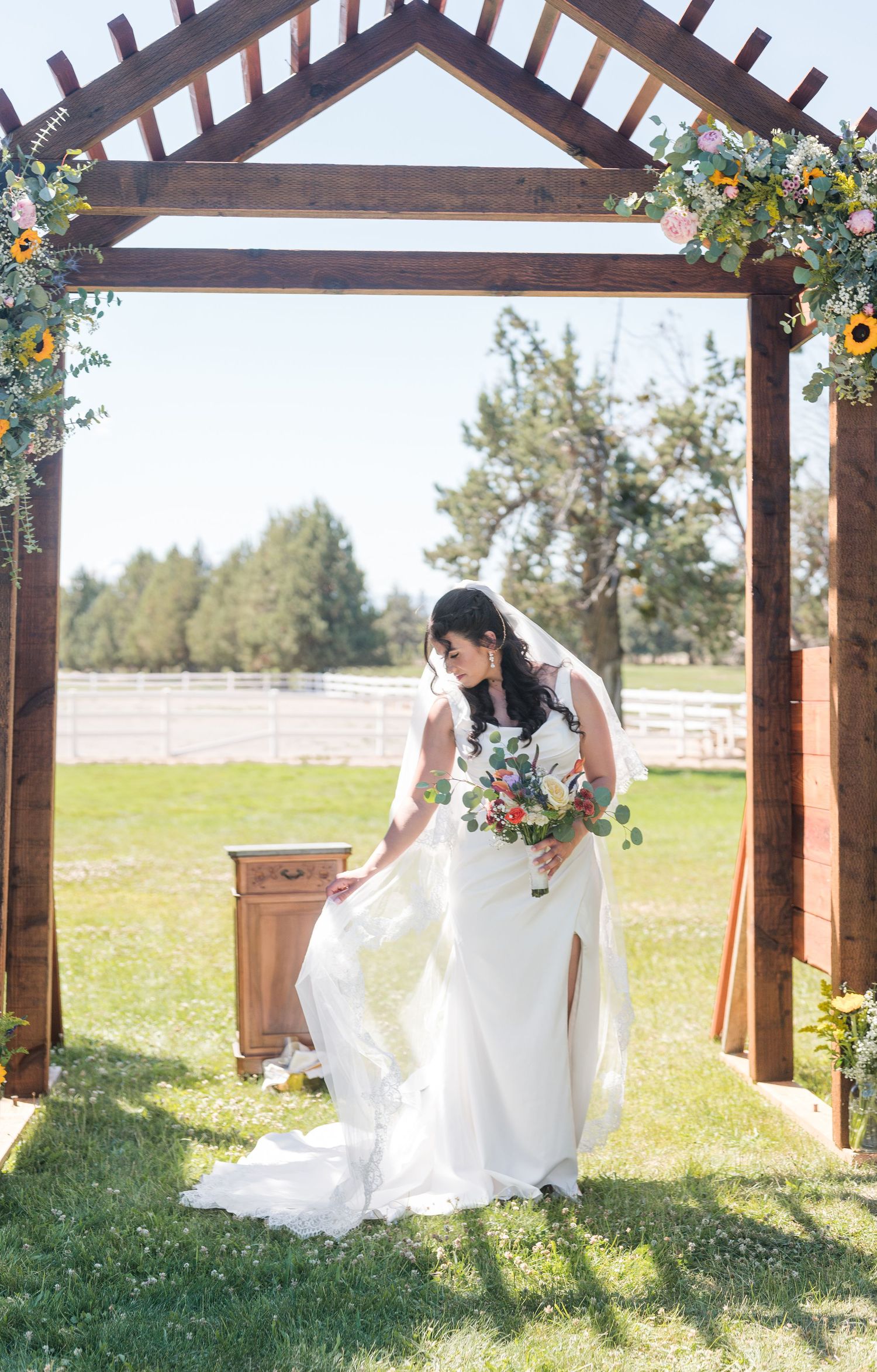 Bride in white gown holding bouquet stands under rustic wooden arch adorned with sunflowers at outdoor wedding.