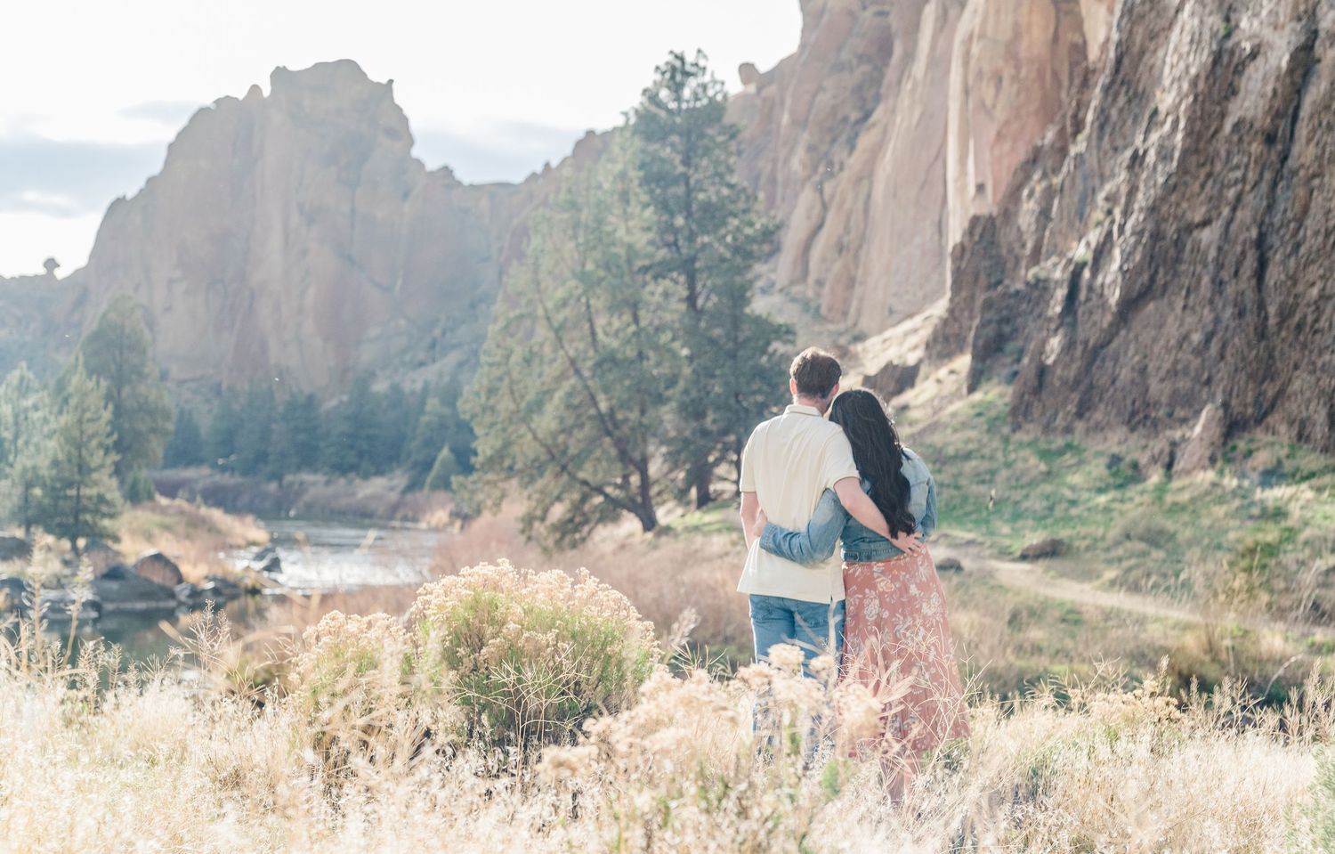 Couple embracing while gazing at scenic rocky cliffs and river landscape in a sunlit outdoor setting.