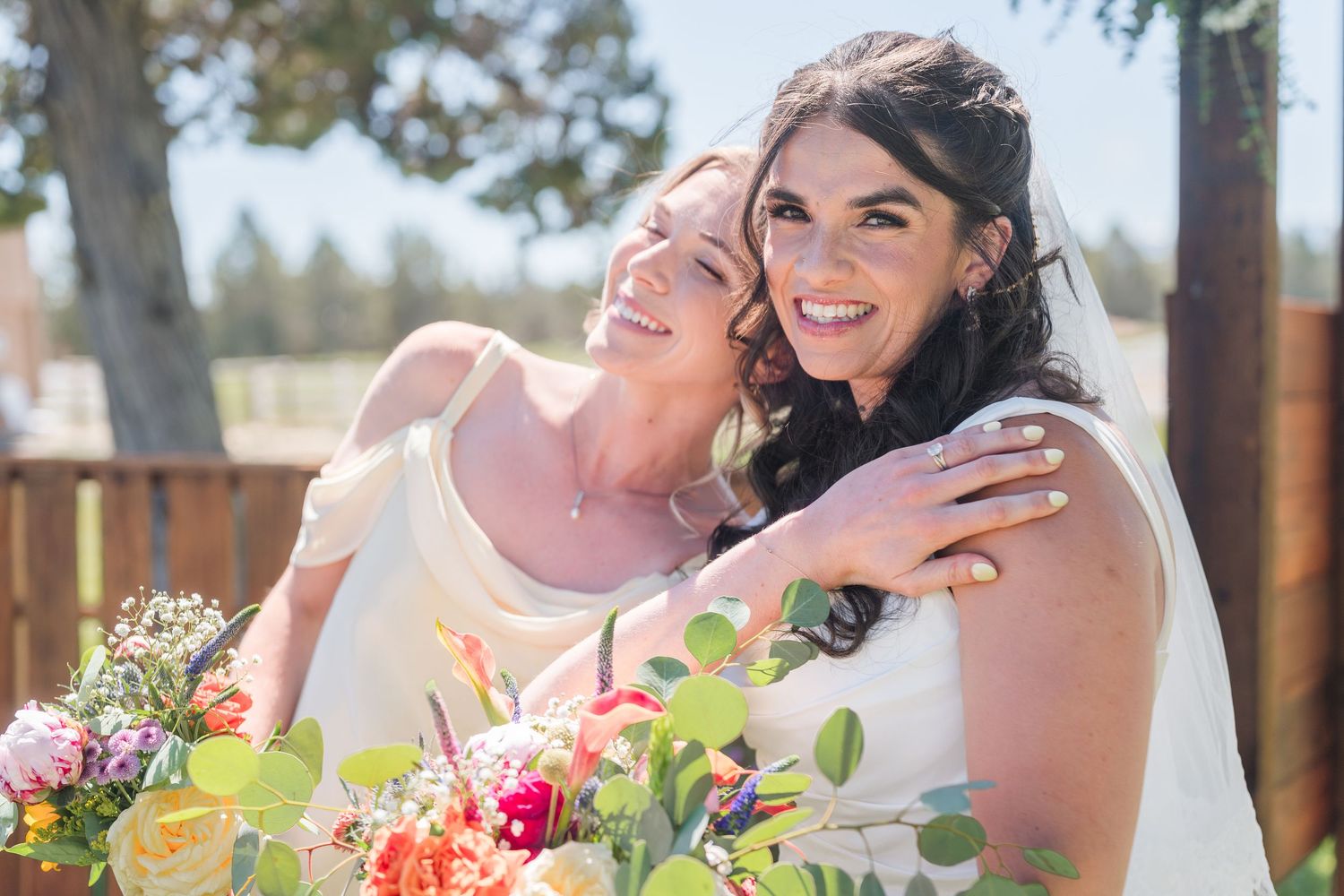 Two smiling brides in white dresses embrace outdoors on a sunny day, holding a colorful bouquet of pink, orange and yellow flowers.