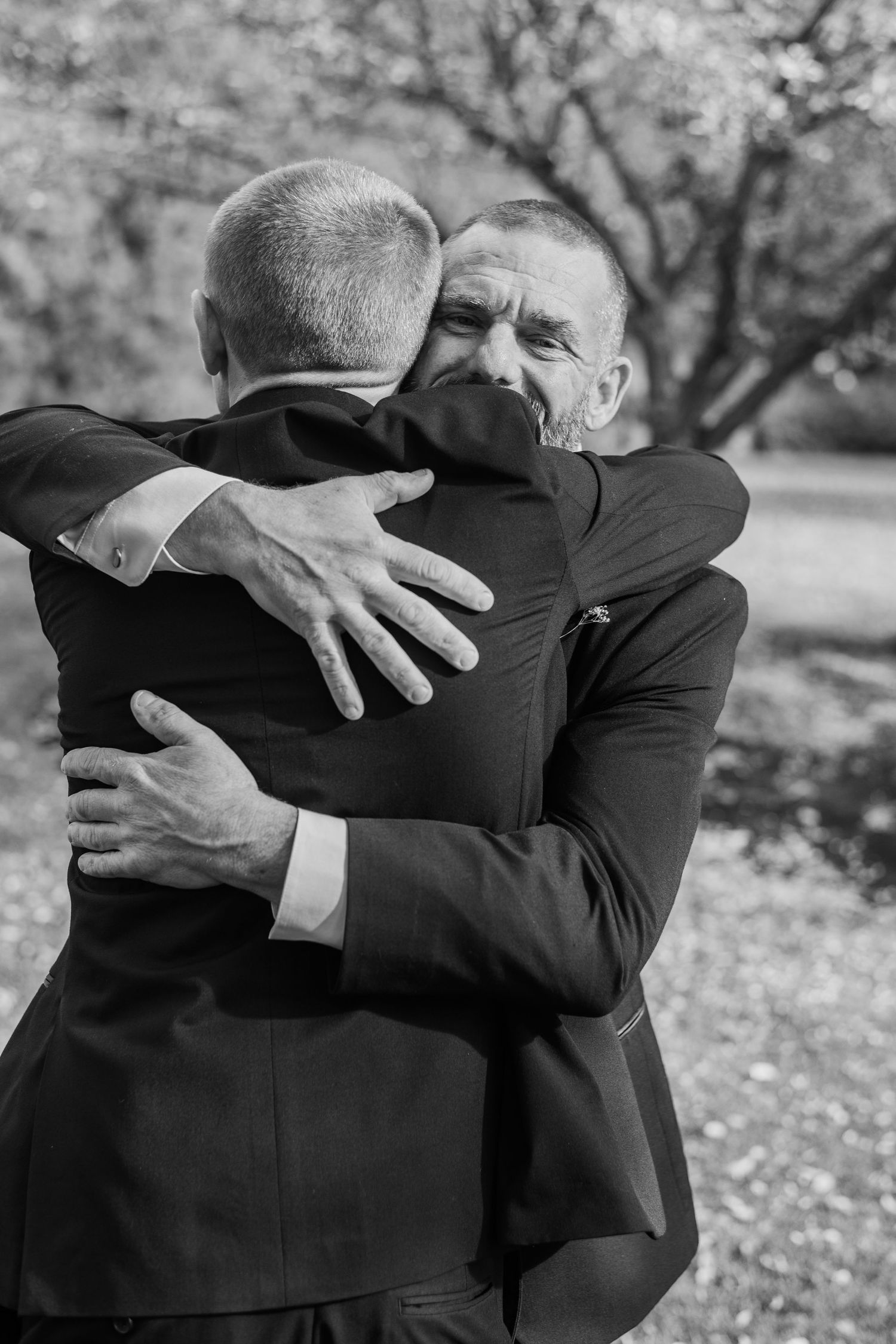 Two men in dark suits share a heartfelt embrace outdoors in a black and white wedding photo.