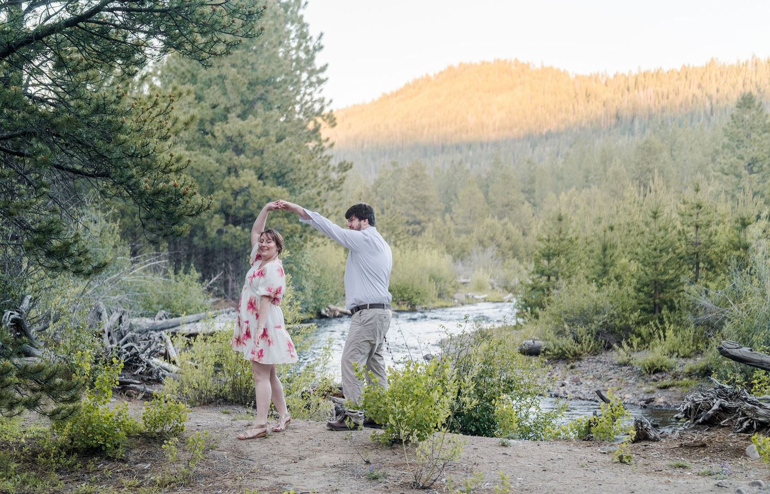 Couple dancing outdoors near a mountain stream surrounded by lush green trees at golden hour.