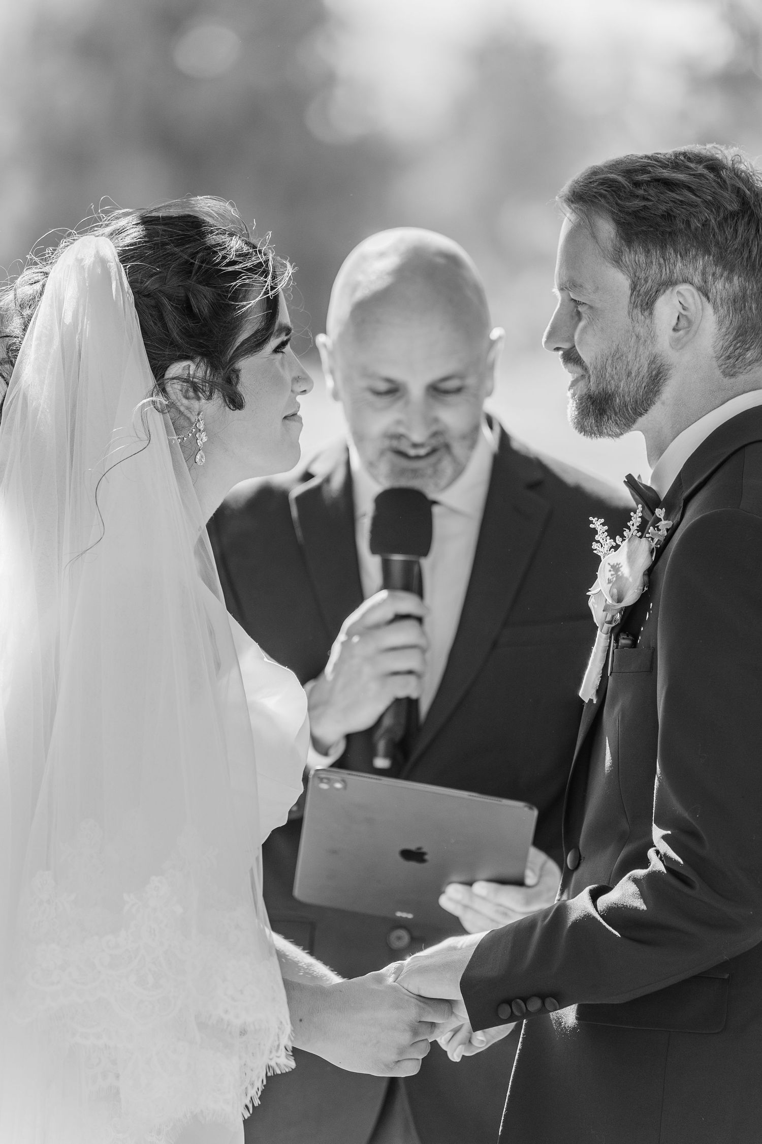 Black and white wedding ceremony photo of bride and groom exchanging vows with officiant holding microphone and iPad.