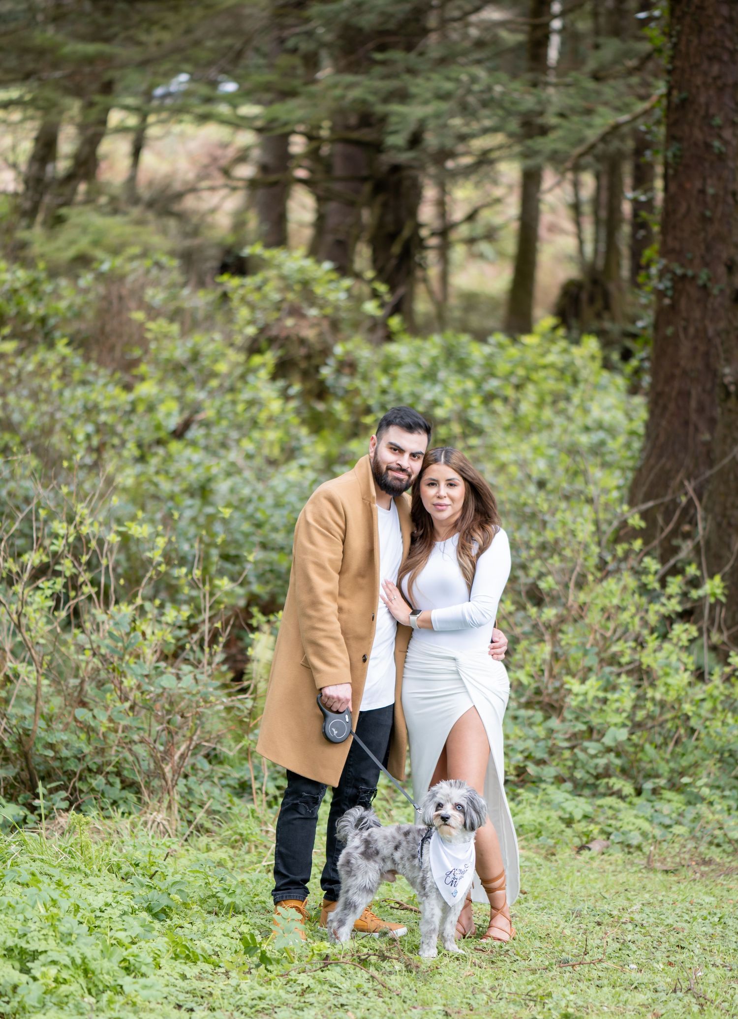 Couple posing with their dog in a lush forest setting, man in tan coat and woman in white dress.