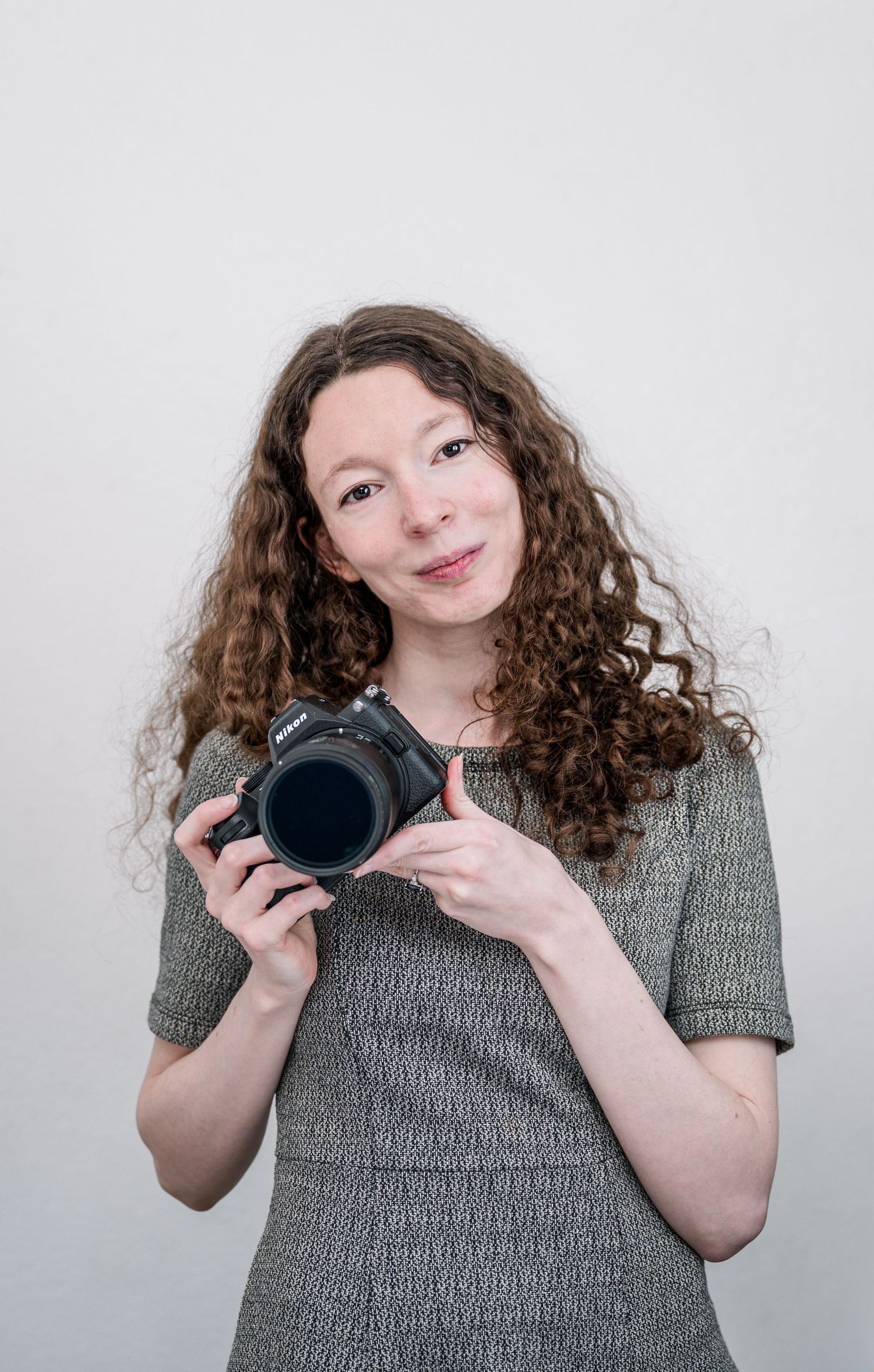 Smiling woman with long curly auburn hair in a gray textured dress holding a black mirrorless camera against a plain light background.