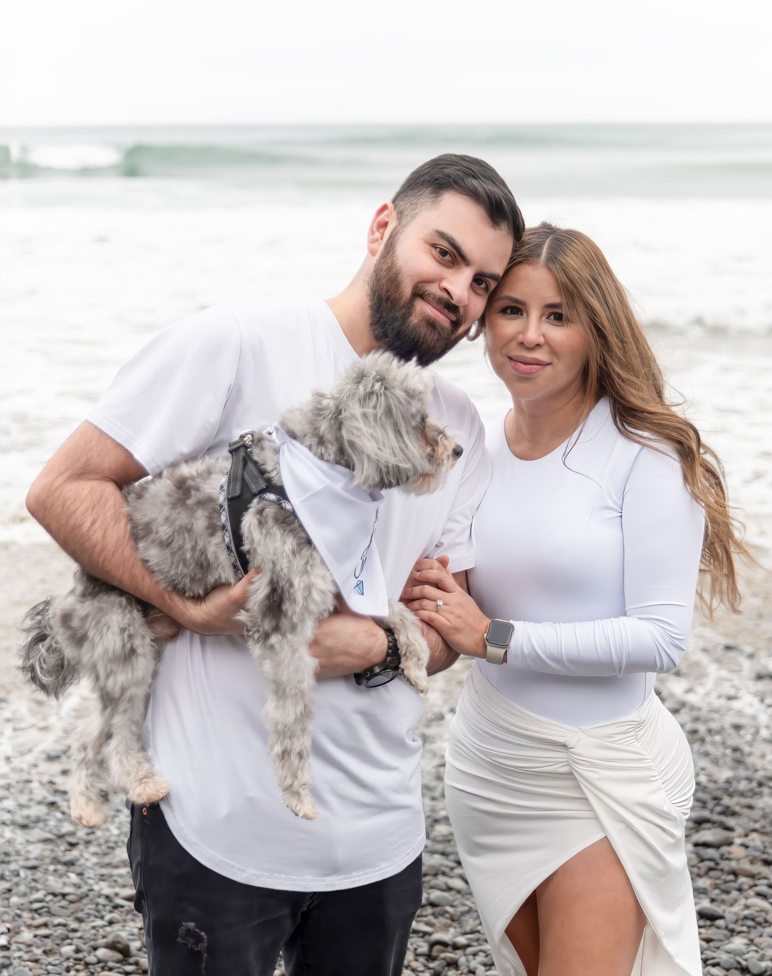Couple in white outfits playing with their fluffy grey dog on pebble beach.