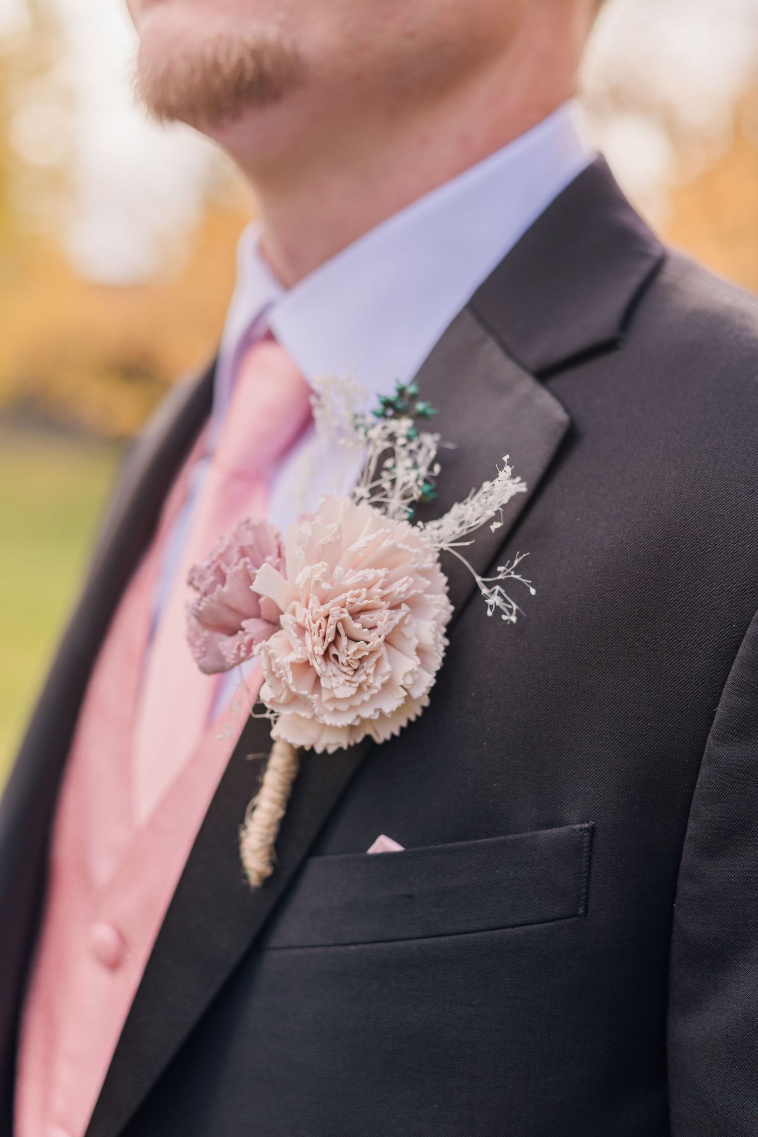 Groom wearing dark suit with pink tie and blush floral boutonniere featuring carnation and baby's breath at wedding.