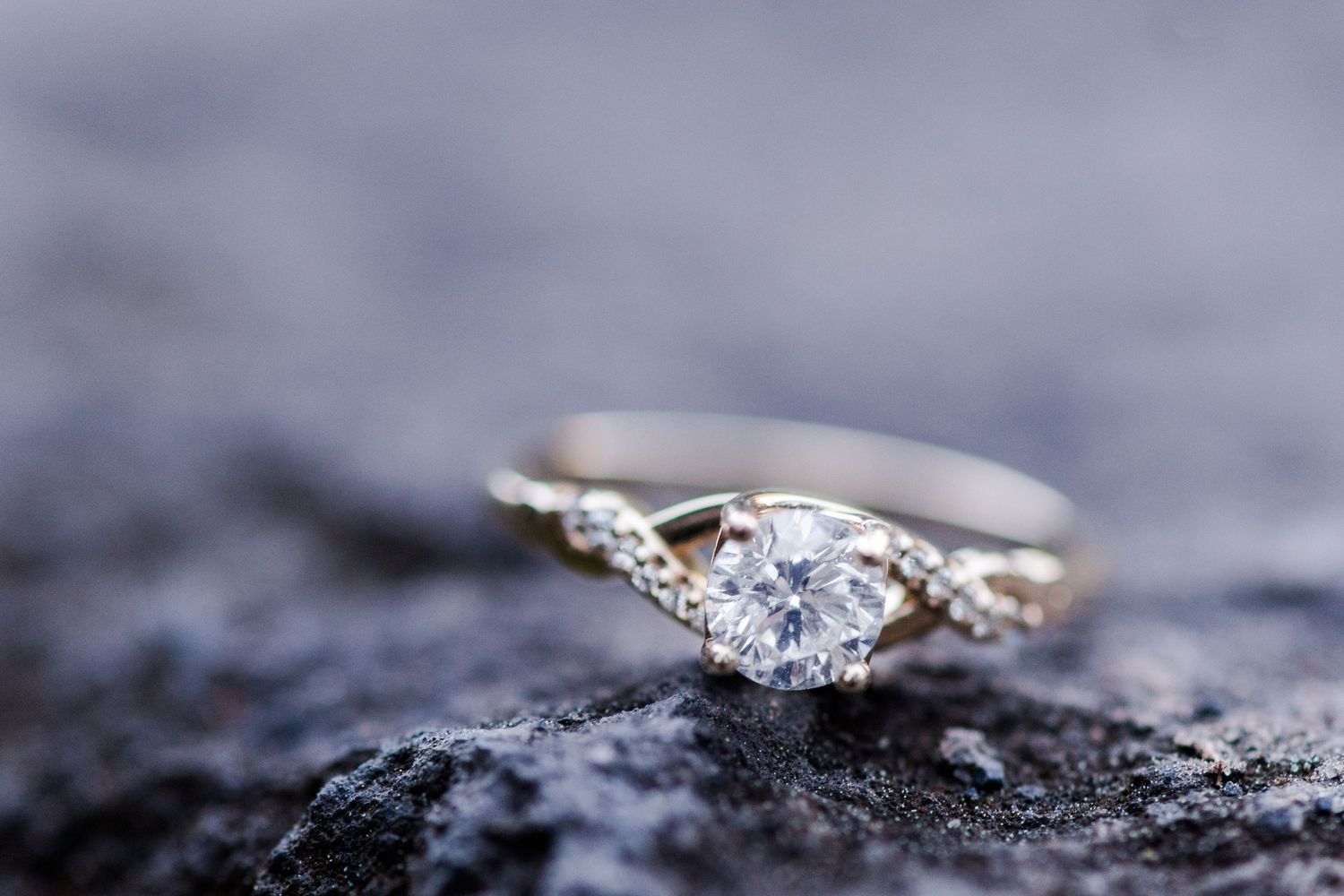Elegant diamond solitaire engagement ring with twisted band resting on dark textured rock surface.
