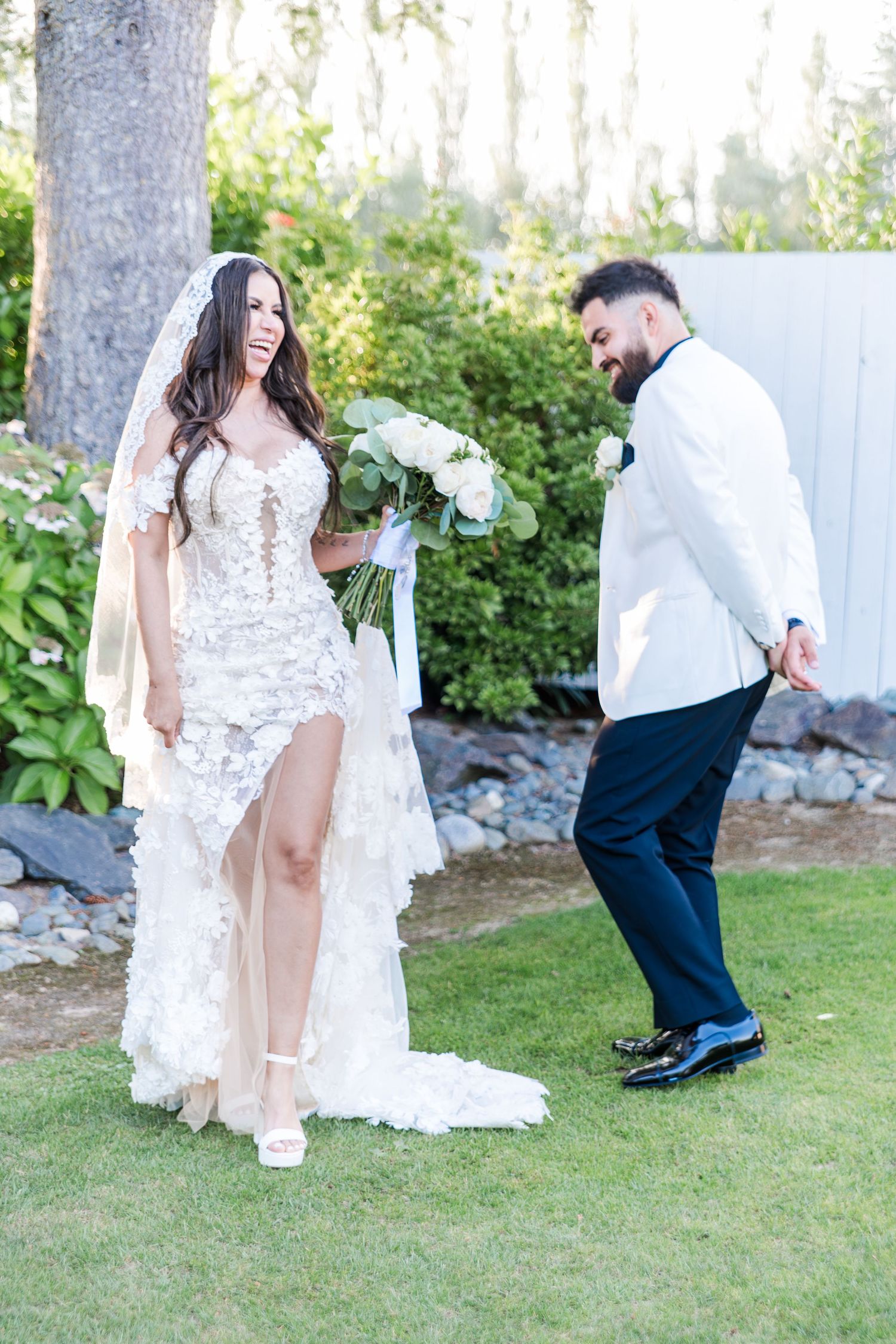 Bride in lace gown with veil holds white bouquet while groom in white jacket bows playfully on garden lawn.