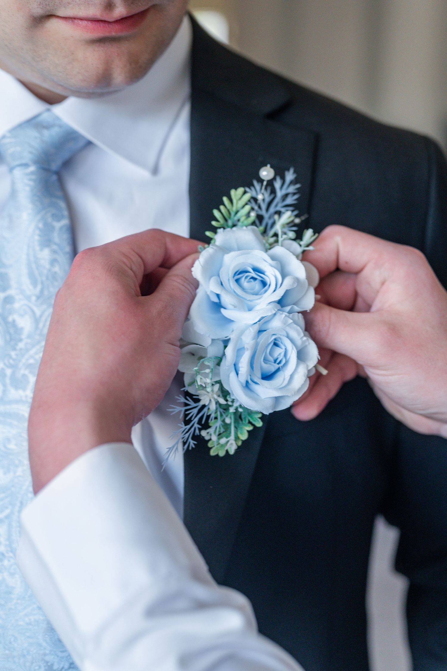 Hands pinning light blue rose boutonniere on groom's dark suit lapel, wedding preparation detail.