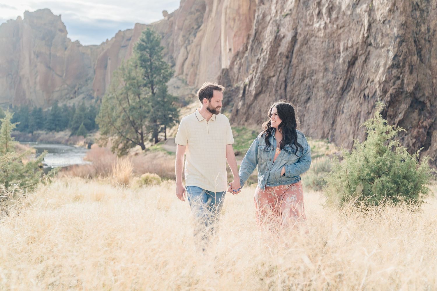 Couple holding hands walking through golden grass fields with dramatic rocky cliffs and river in background.