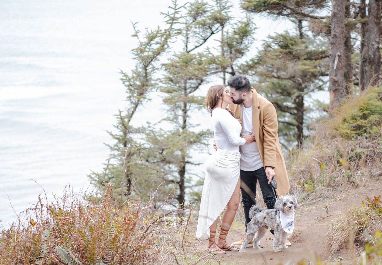 Couple kissing on a coastal cliff trail with two dogs, surrounded by pine trees and ocean views.